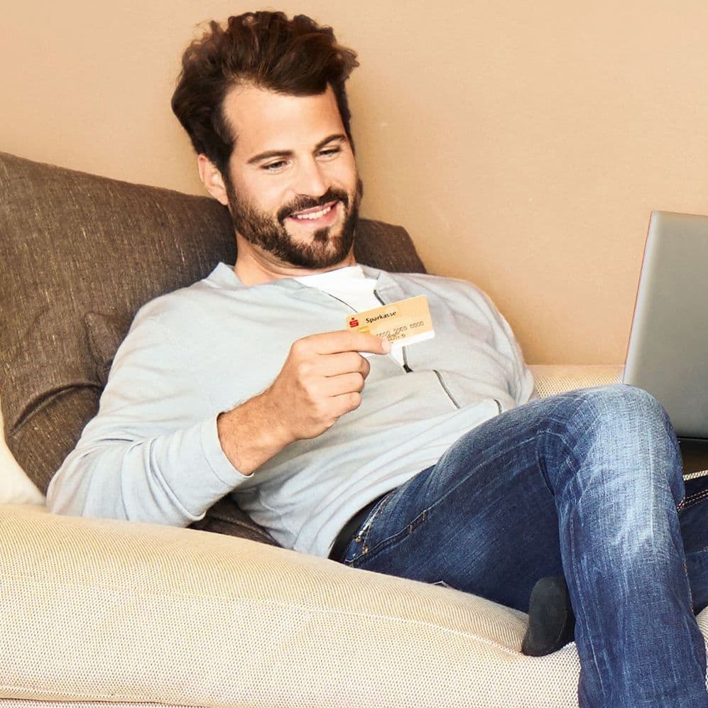 Mann sitzt mit einer Kreditkarte und seinem Laptop auf dem Schoß auf der Couch.
