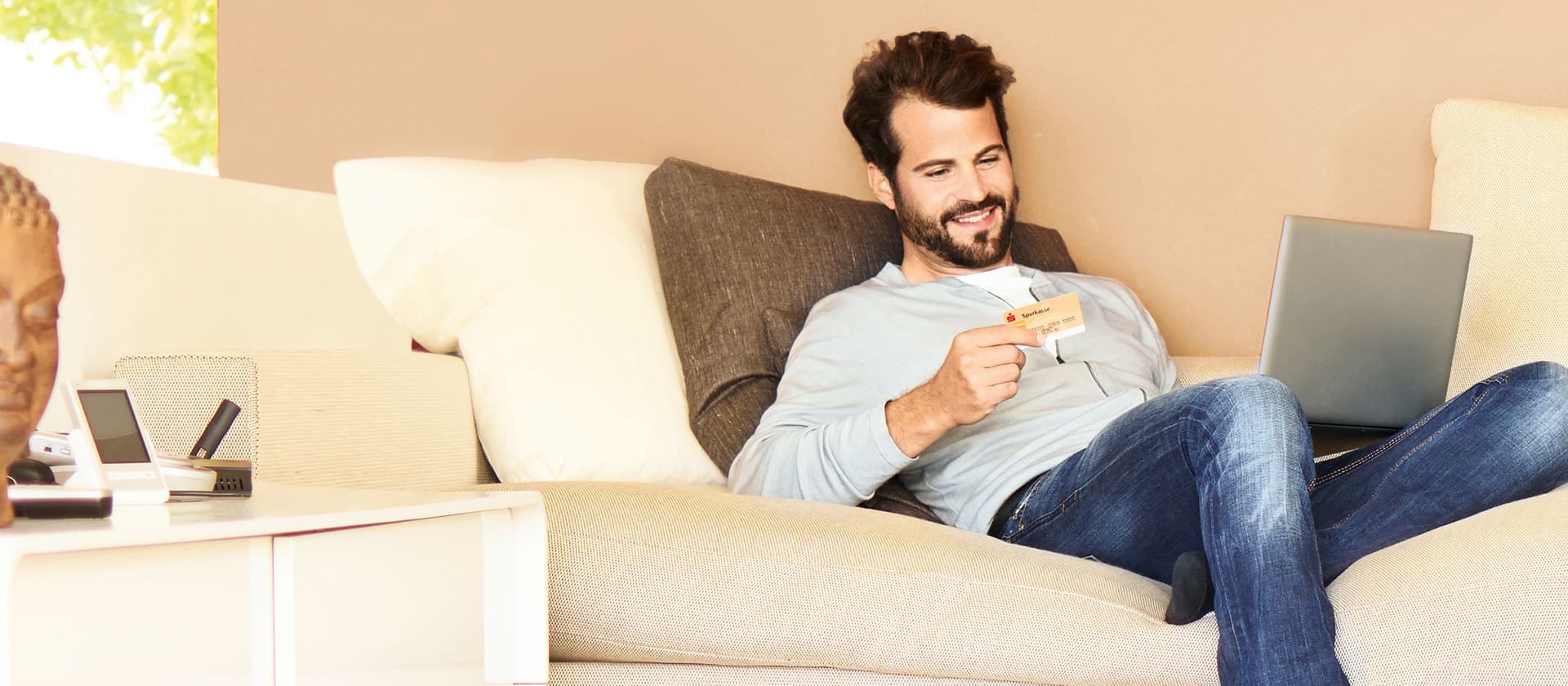Mann sitzt mit einer Kreditkarte und seinem Laptop auf dem Schoß auf der Couch.