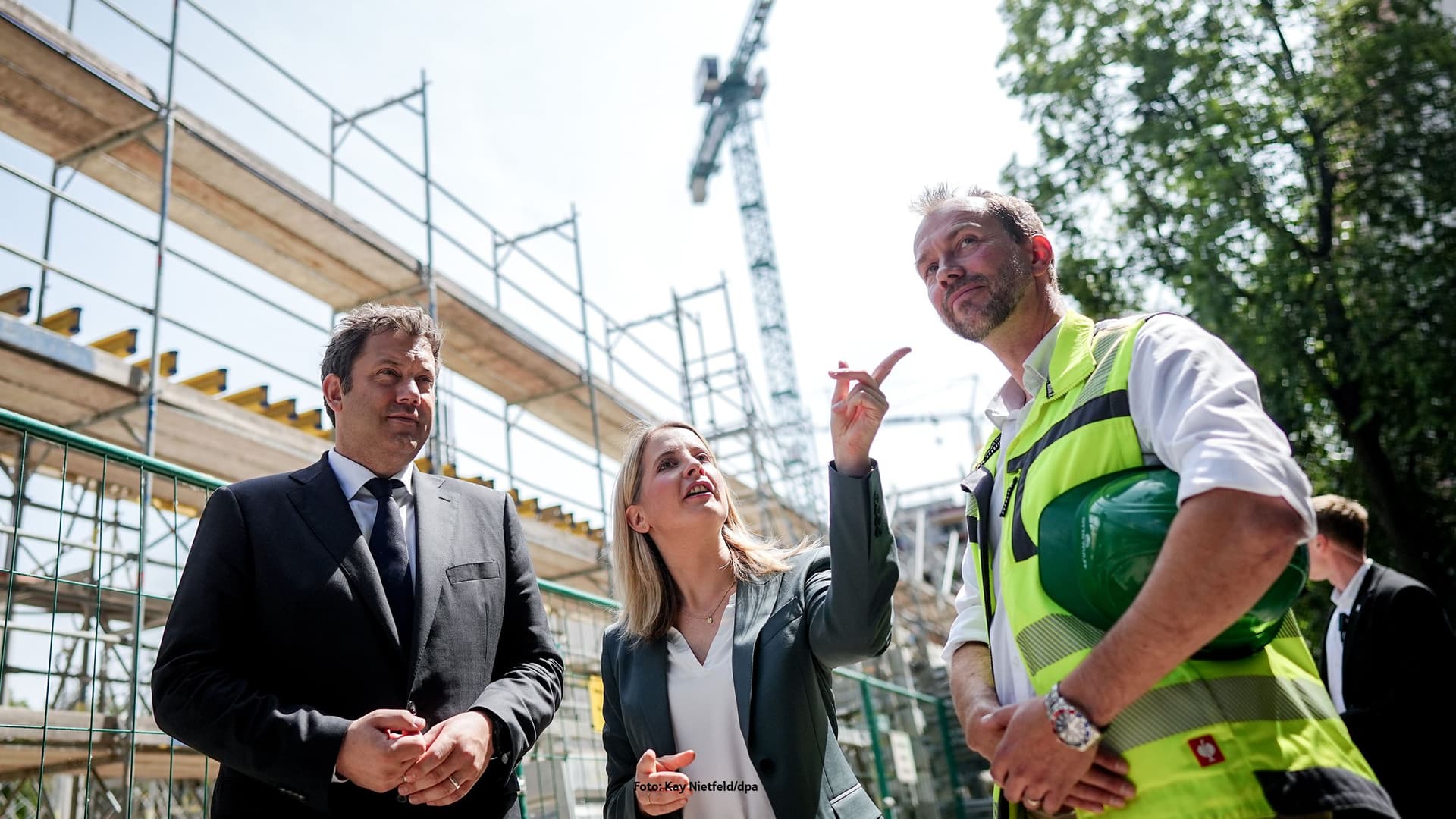 Drei Personen auf einer Baustelle, zwei in Businesskleidung und eine in Sicherheitsweste mit Helm, im Hintergrund ein Baugerüst und ein Kran