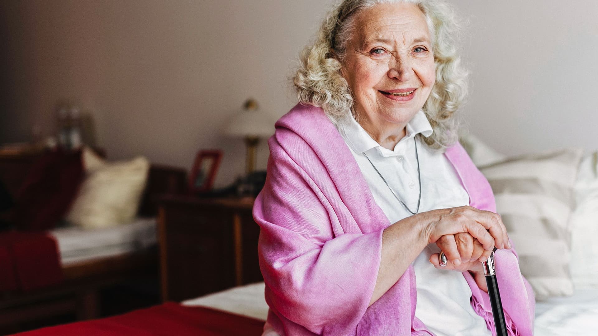 Eine ältere Frau sitzt mit ihrem Gehstock auf einem Bett und lächelt in die Kamera.