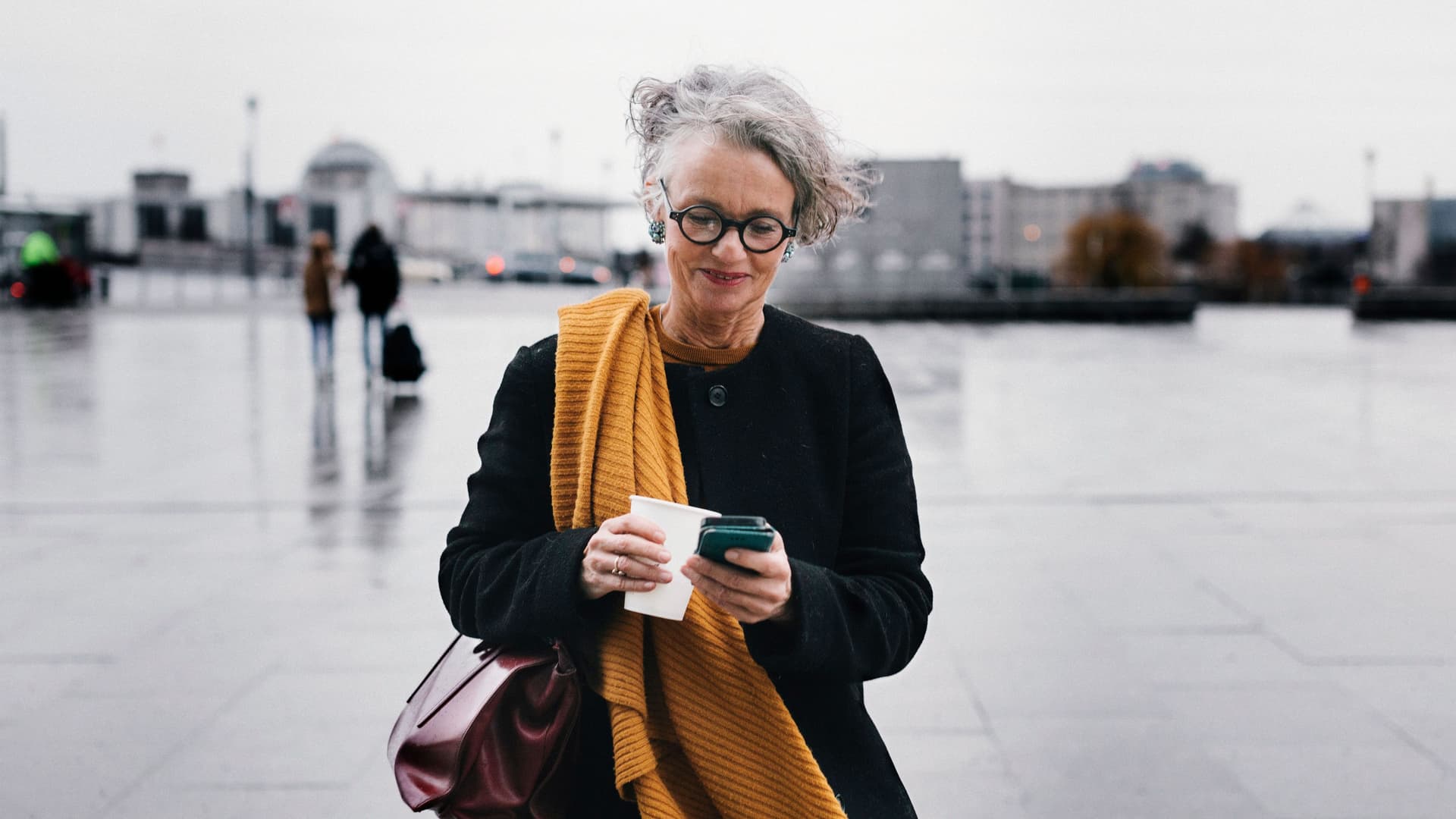 Eine ältere Frau steht mit einem Smartphone und einem Kaffeebecher in der Hand auf einem Platz. Sie schreibt und guckt auf ihr Handy.