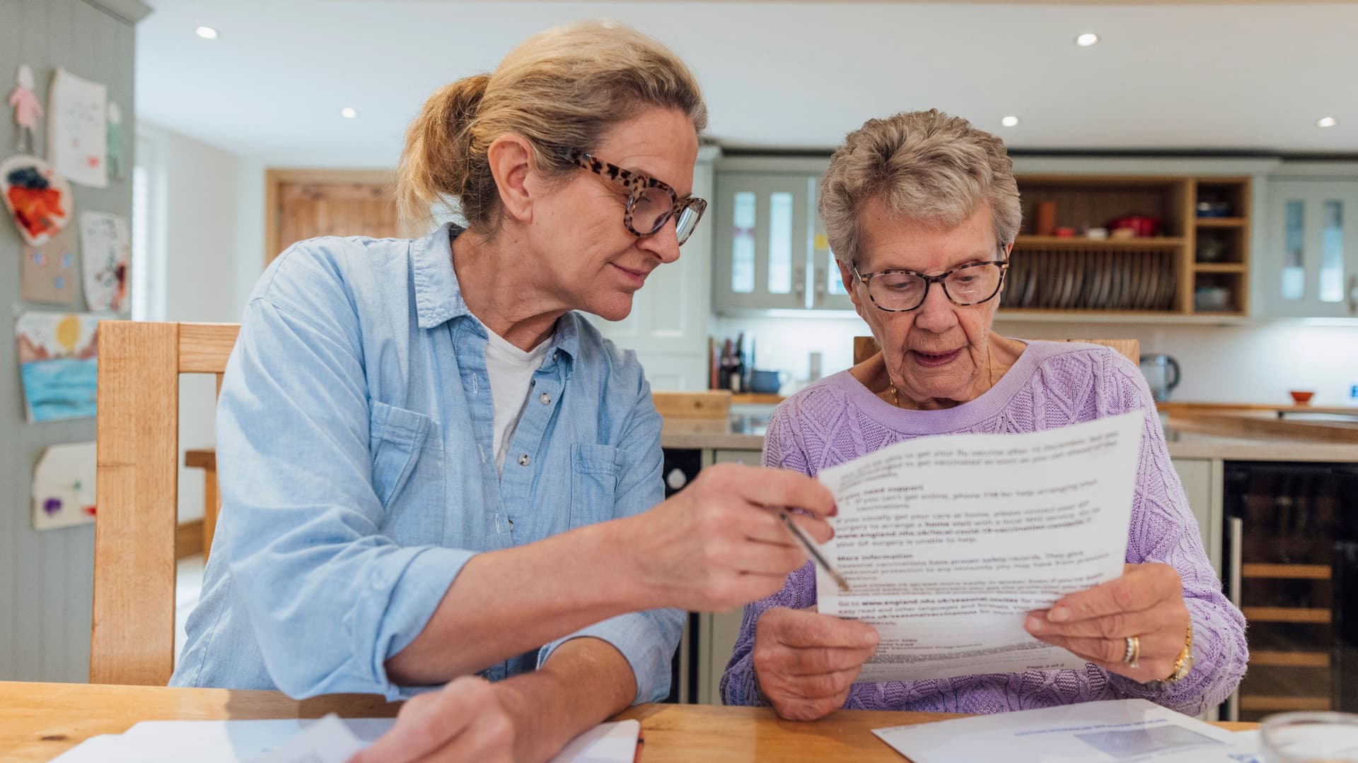 Eine ältere Frau und ihre erwachsene Tochter sitzen am Esstisch und schauen besorgt Finanzrechnungen durch.