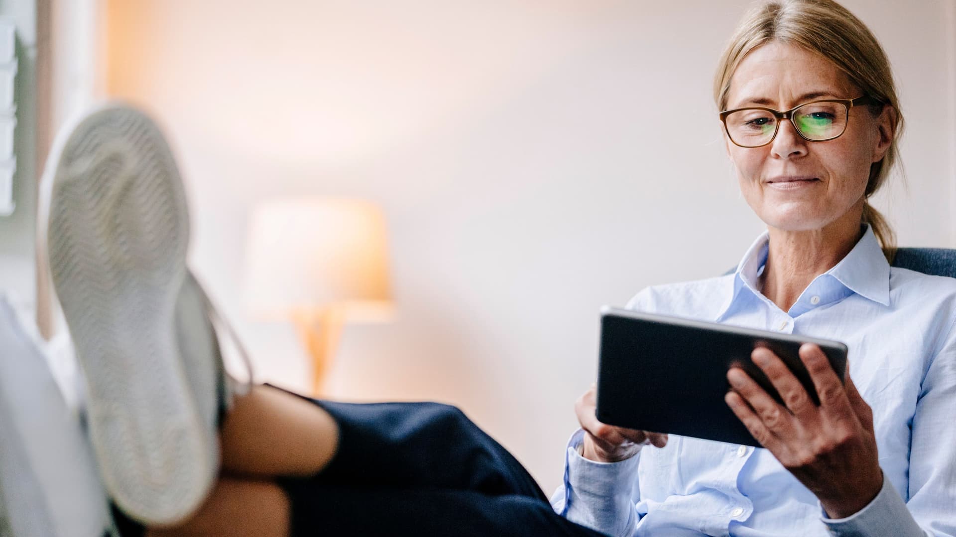 Eine Frau mittleren Alters hält ein Tablet in der Hand und legt entspannt ihre Füße auf den Tisch.