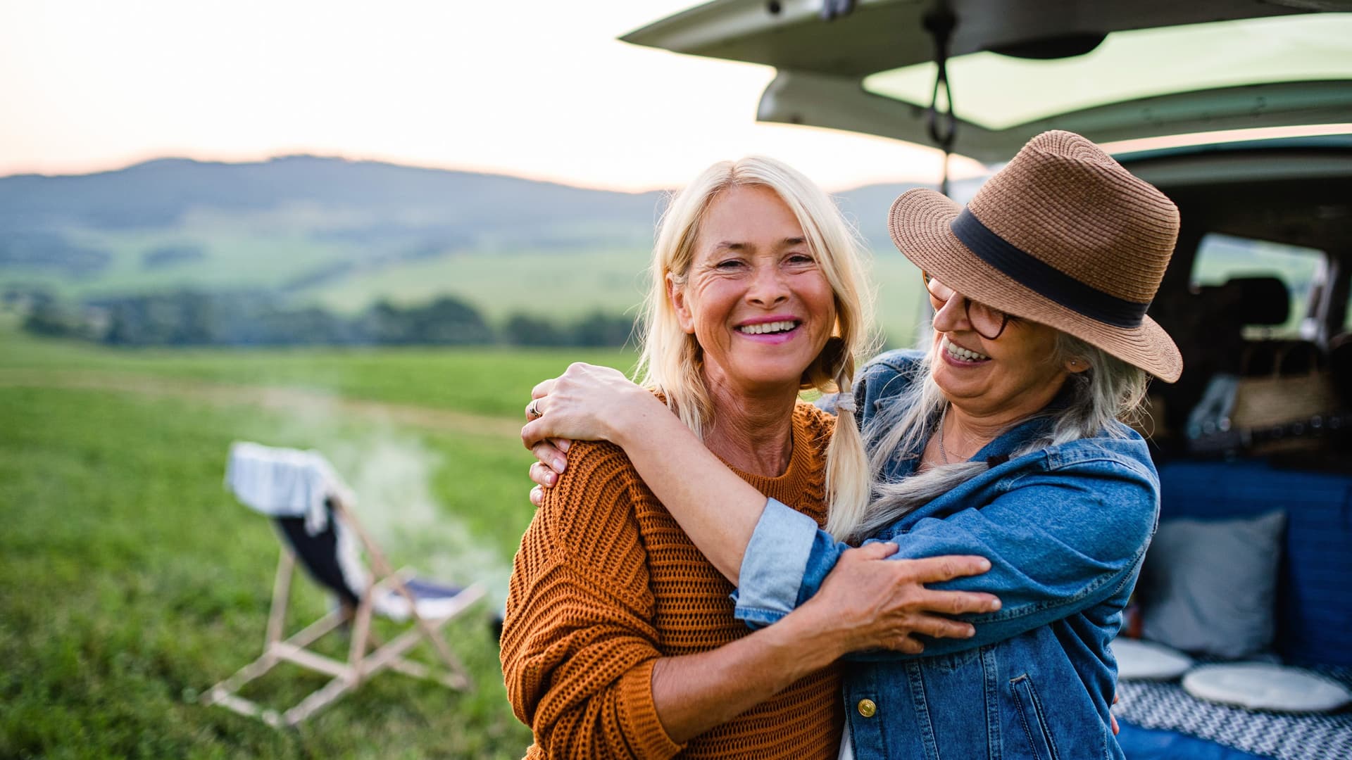 Ältere Freundinnen, die draußen in der Natur in die Kamera blicken und sich vor einem Van lachend umarmen.