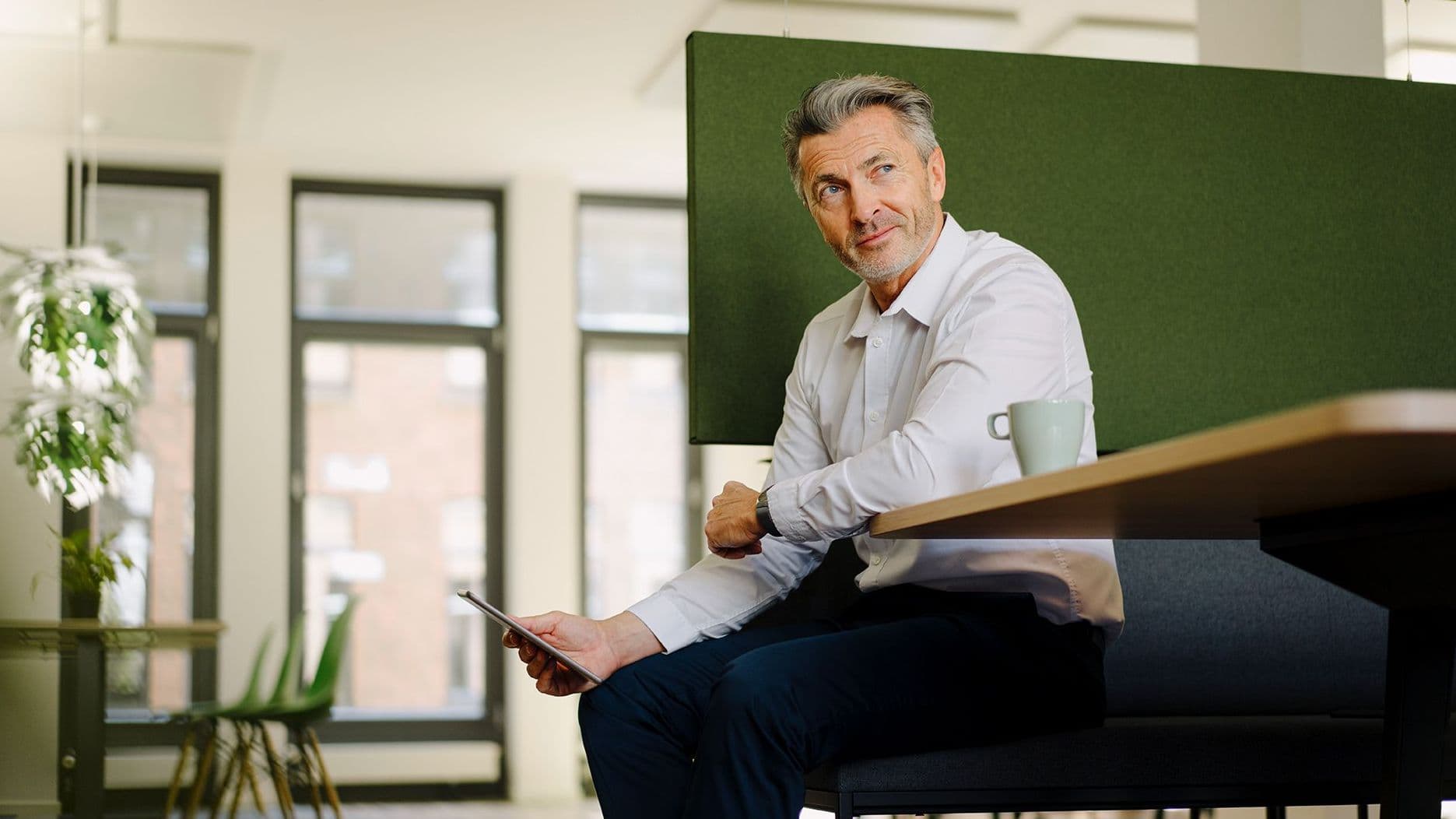 Älterer Mann sitzt in einem großen Raum auf einer Sitzgelegenheit und hat ein Tablet in der Hand. Er schaut zur Seite.
