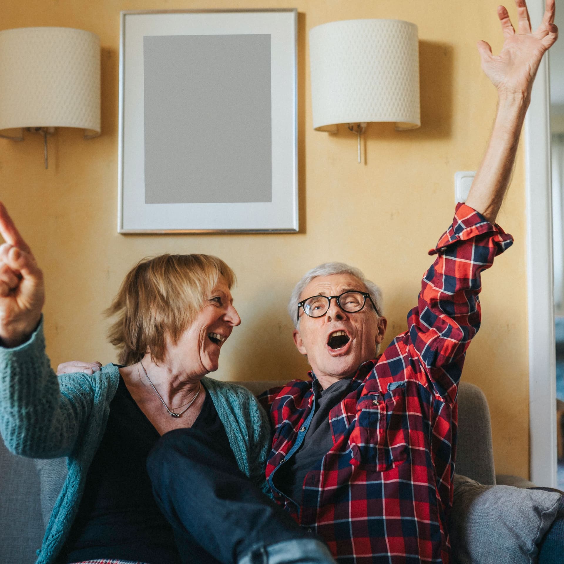 Eine ältere Frau und ein älterer Mann sitzen auf einem Sofa und heben mit fröhlichem Gesichtsausdruck die Hände.
