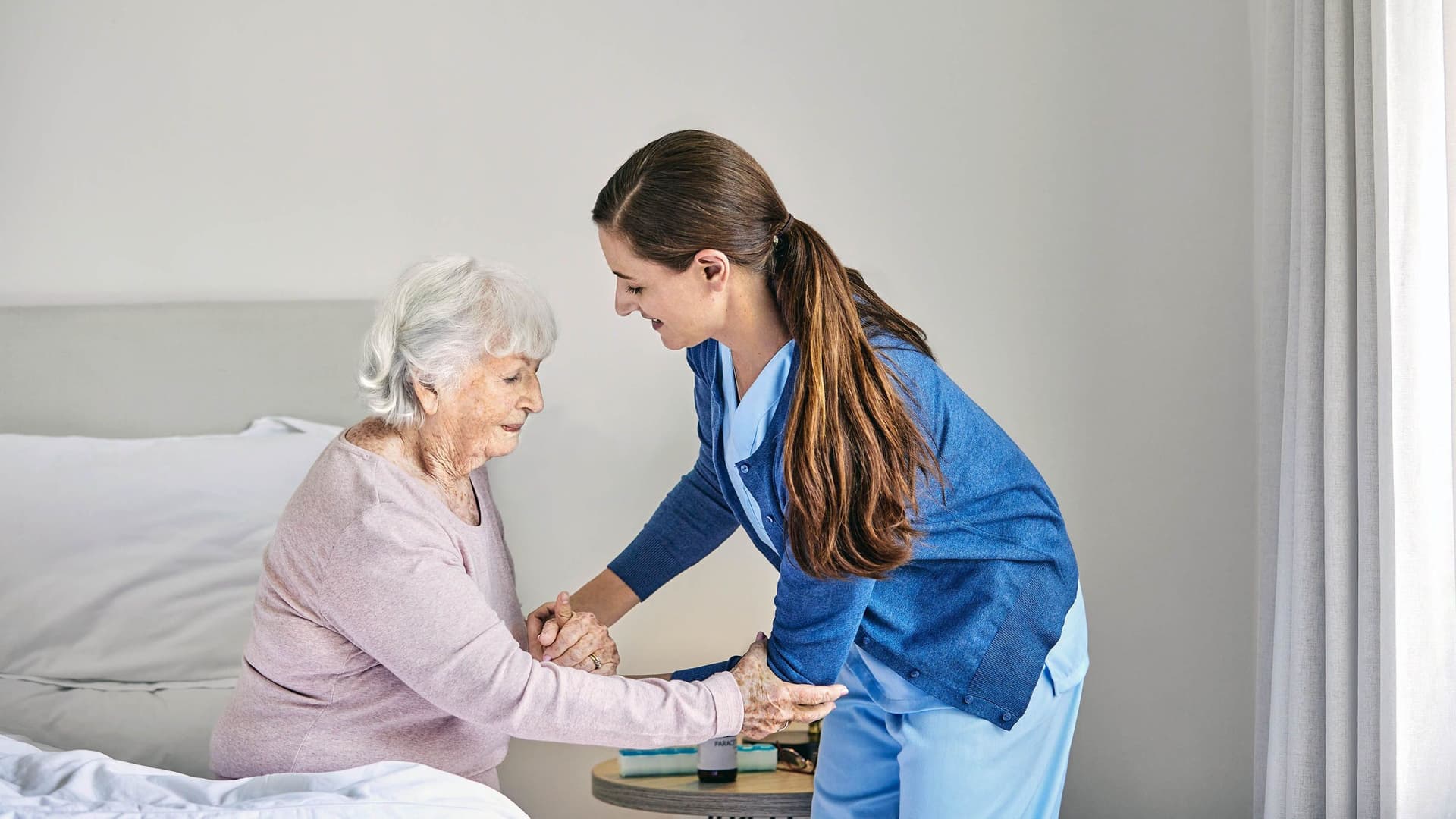 Eine junge Pflegerin hilft einer älteren Frau, die am Bettrand sitzt, beim Aufstehen. Sie hält die Seniorin unterstützend an den Armen. Der Raum ist hell und schlicht eingerichtet.