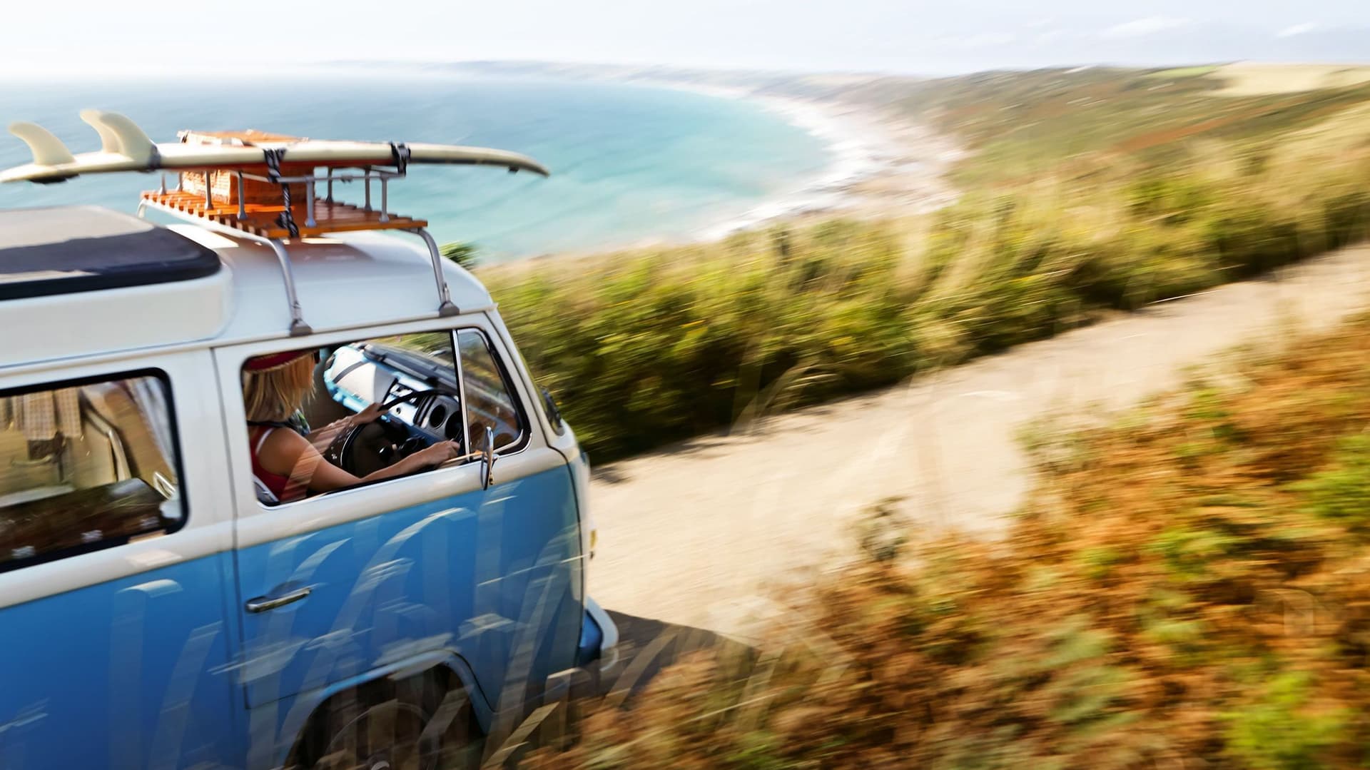 Blauer Bulli mit Surfbrett fährt auf einer sommerlichen Küstenstraße entlang