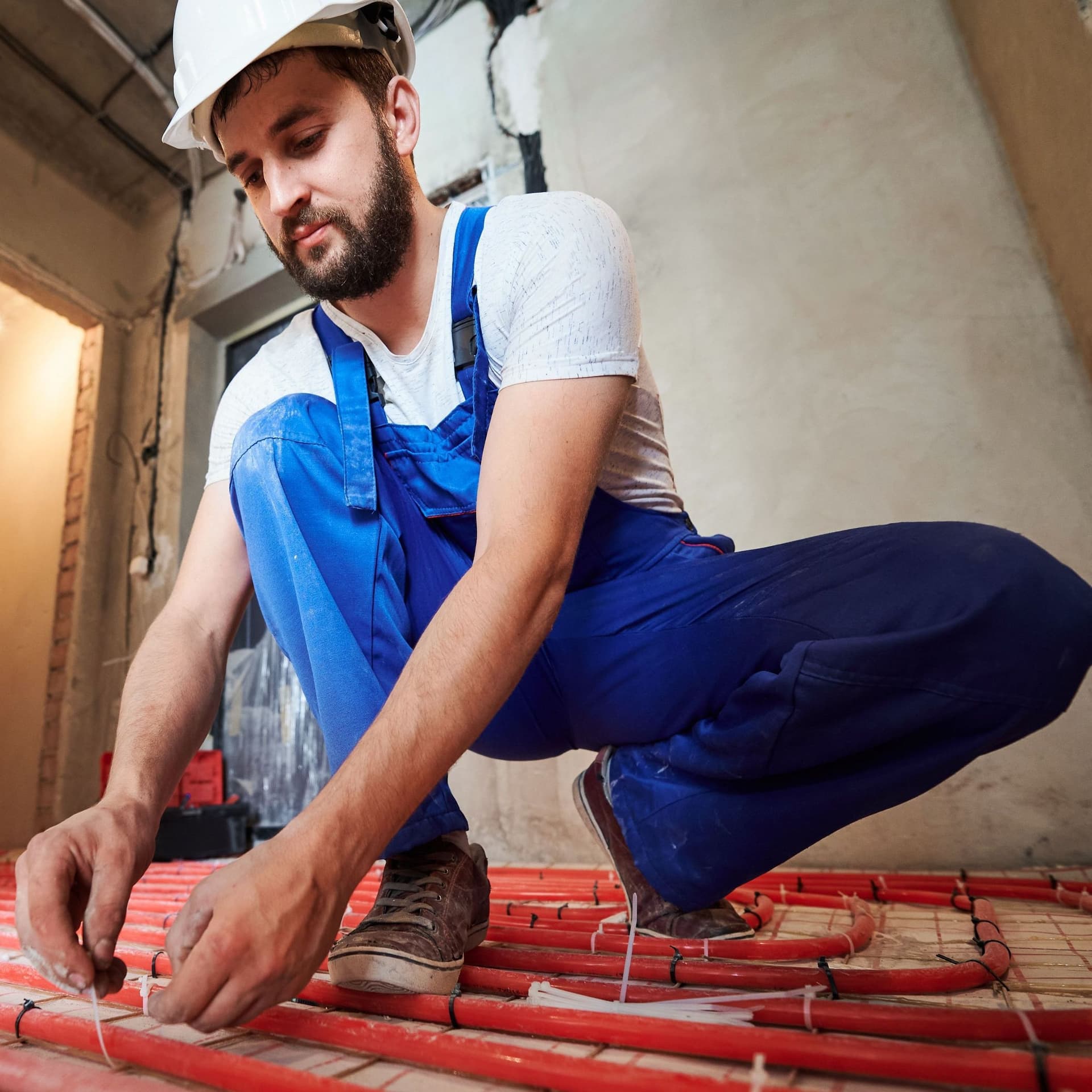 Mann mit blauer Arbeitslatzhose und weißem Schutzhelm installiert eine Fußbodenheizung.