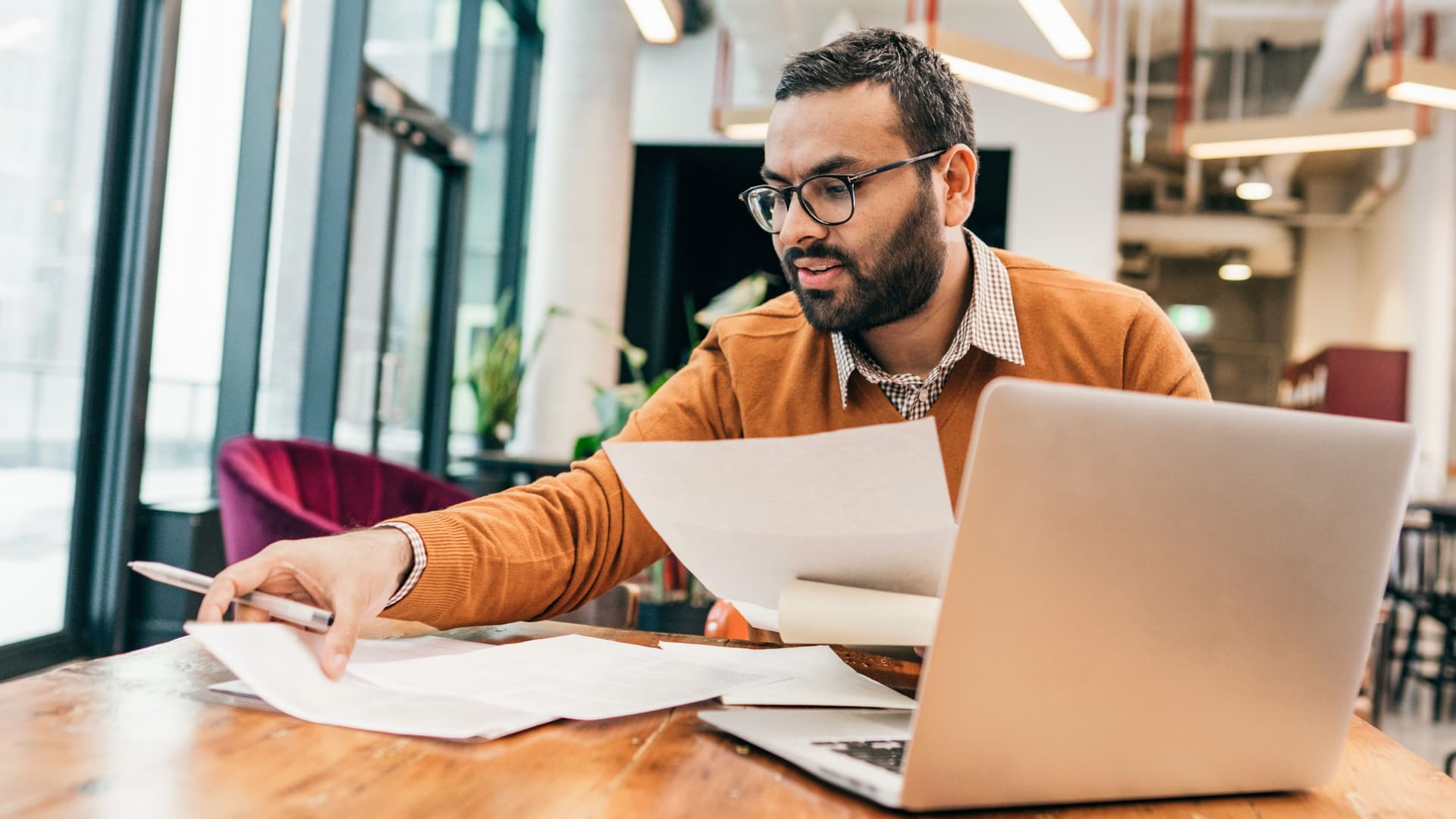 Mann mit Bart und Brille sitzt an einem Tisch mit aufgeklapptem Laptop und hält mehrere Blatt Papier und einen Stift in den Händen.