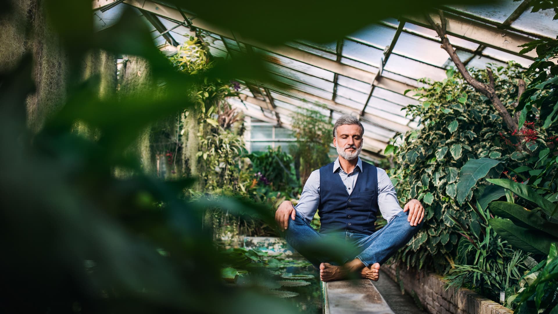 Ein Mann sitzt barfüßig im Schneidersitz auf einer Mauer in einem Gewächshaus mit vielen Pflanzen. Er hat die Augen geschlossen und trägt schicke Kleidung.
