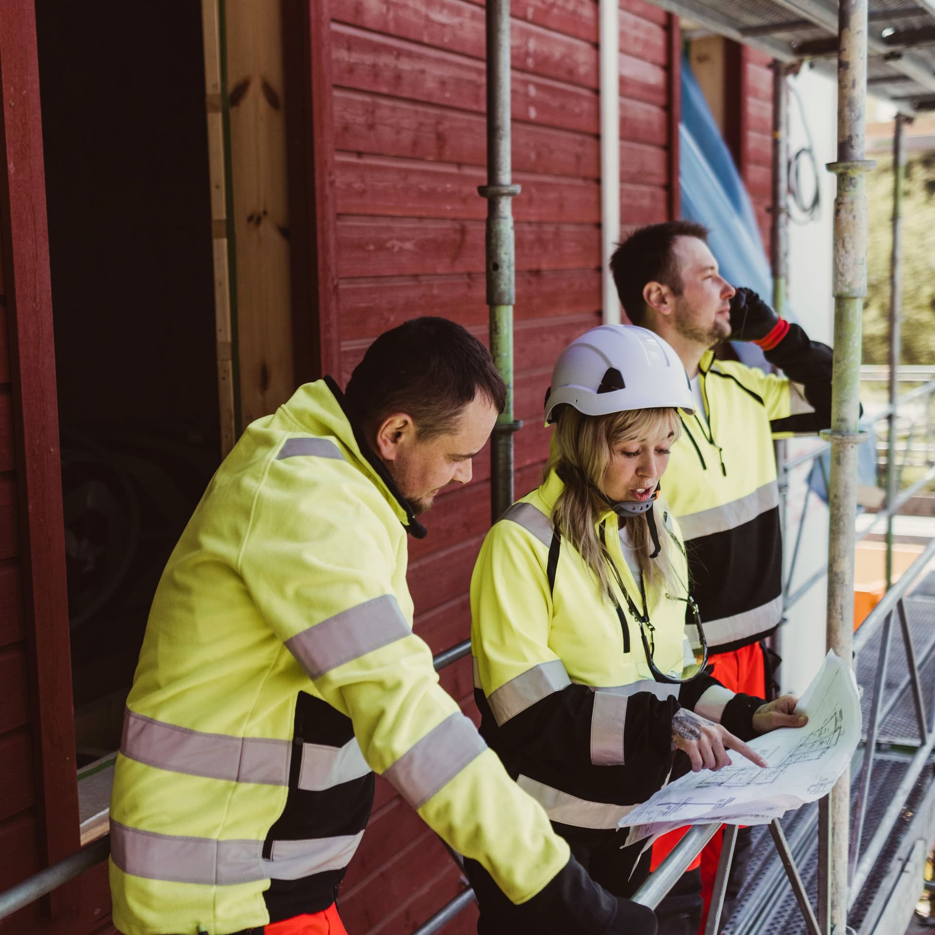 Eine Frau und ein Mann in gelber Arbeitsbekleidung besprechen einen Bauplan. Im Hintergrund ist ein weiterer Mann in gelber Arbeitskleidung telefonierend zu erkennen.