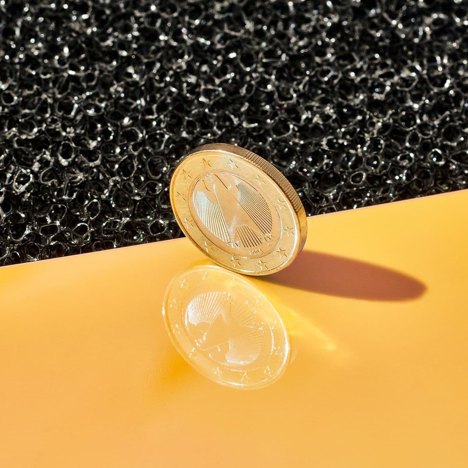Eine Ein-Euro-Münze steht hochkant an der Kante einer gelben, spiegelnden Untergrundfläche.