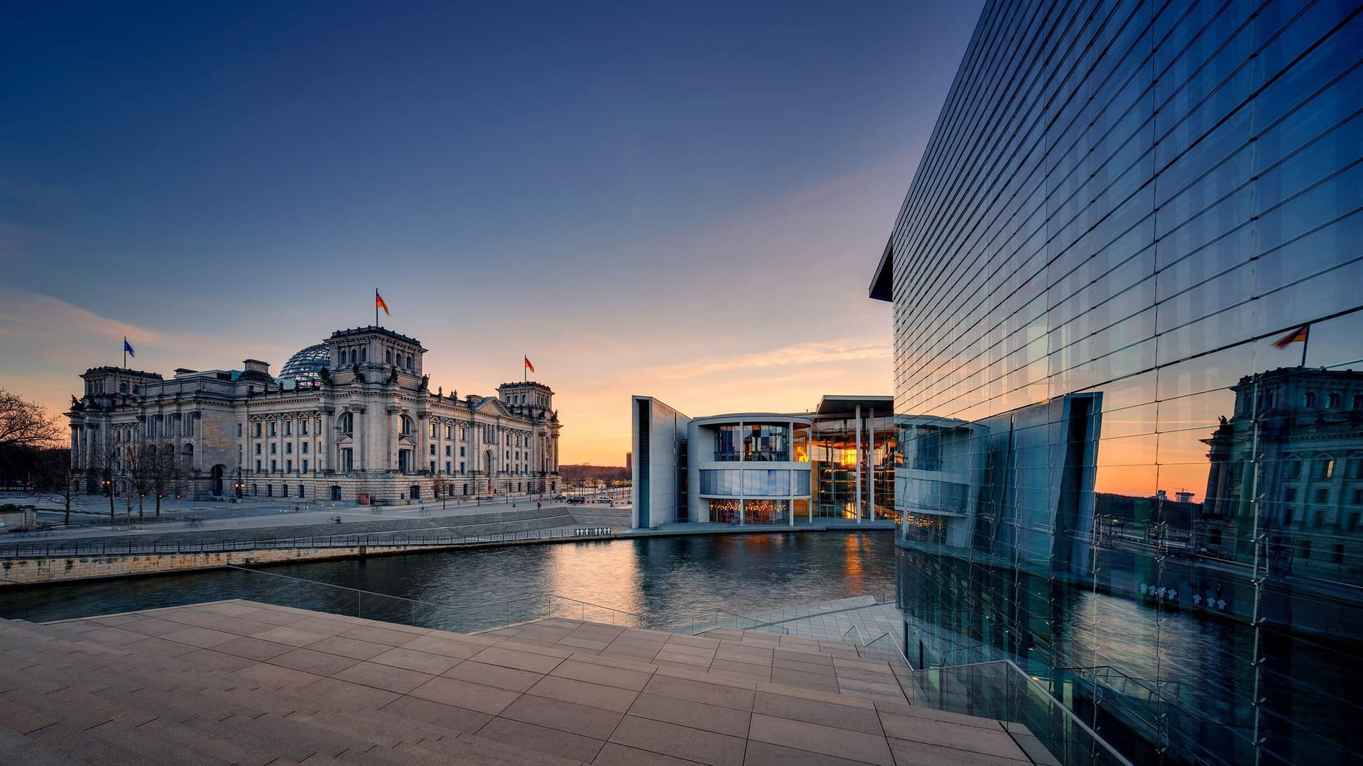 Blick auf den Reichstag mit dem Marie-Elisabeth-Lüders-Haus im Vordergrund. Es dämmert und die Regierungsbauten sind in ein blaues und rotes Licht getaucht.