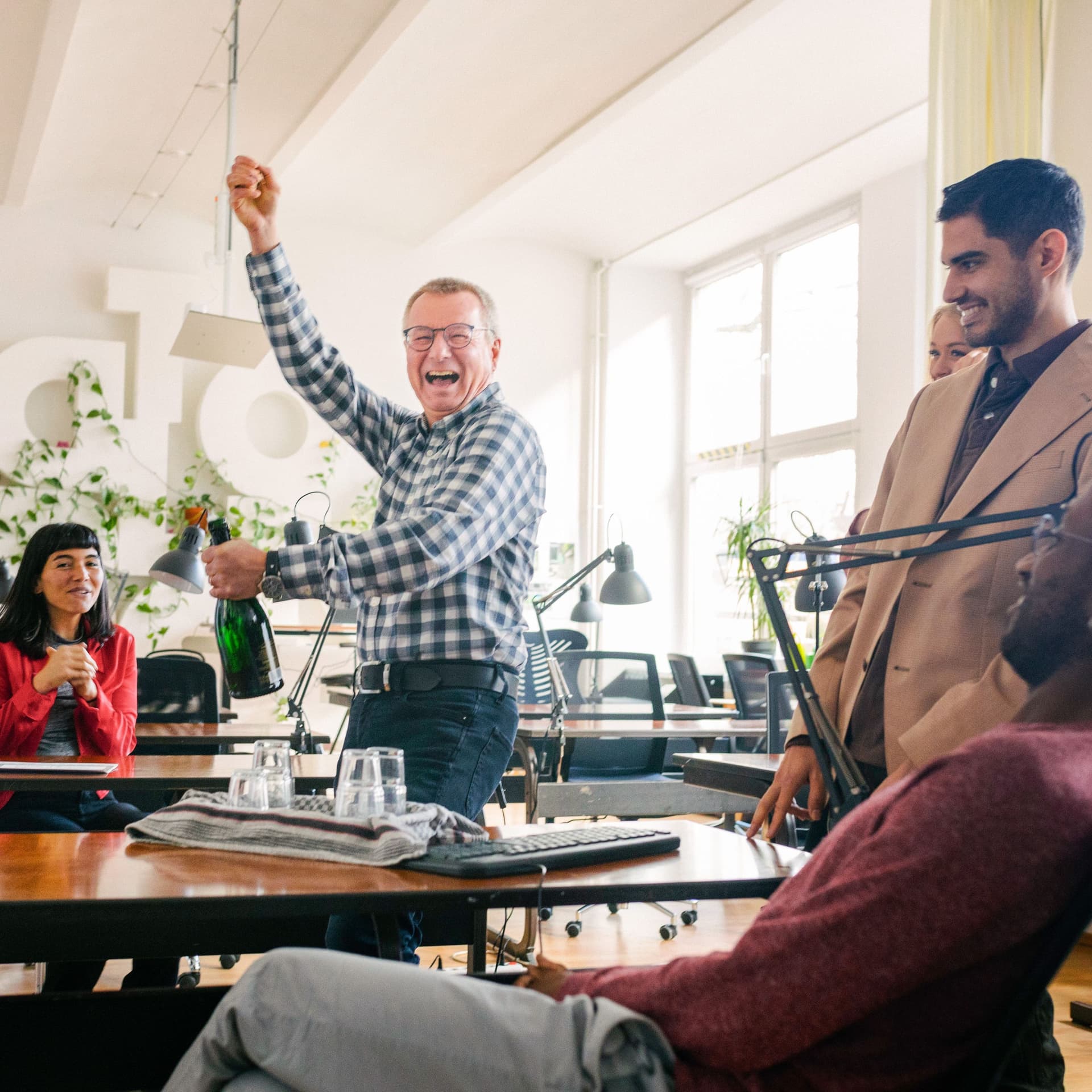 Eine diverse Personengruppe an Schreibtischen. Ein Mann hält eine Sektflasche und lacht.