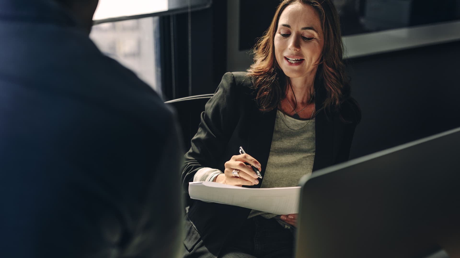 Blick über die Schulter eines Mannes in einem Büro. Vor ihm sitzt eine Frau, die über Papiere in ihrer Hand spricht.