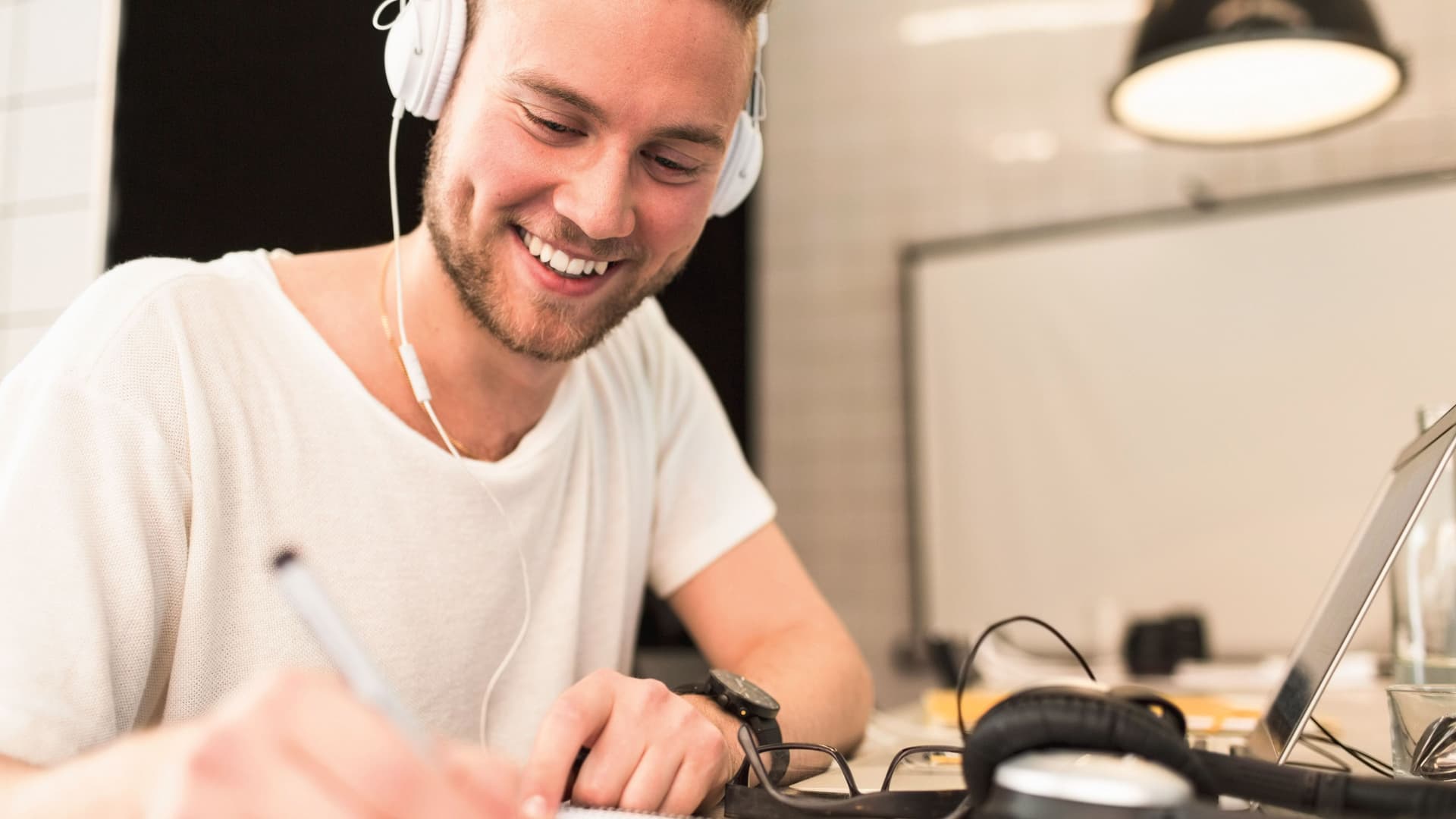 Close-up eines jungen Mannes, der lächelnd an einem Schreibtisch neben einem Laptop sitzt und etwas auf ein Papier notiert.