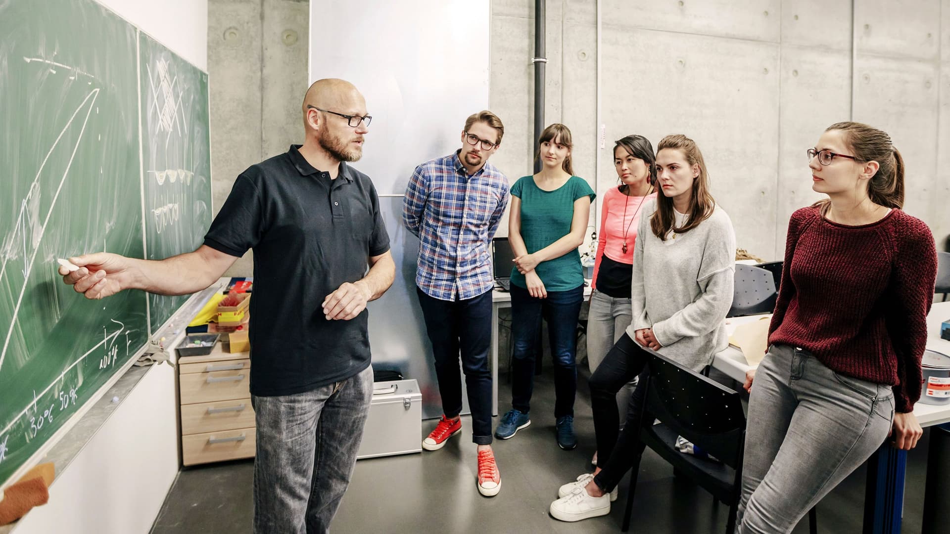 Studenten stehen vor einer Tafel und hören ihrem Lehrenden zu, der eine Kurve an die Tafel gemalt hat.