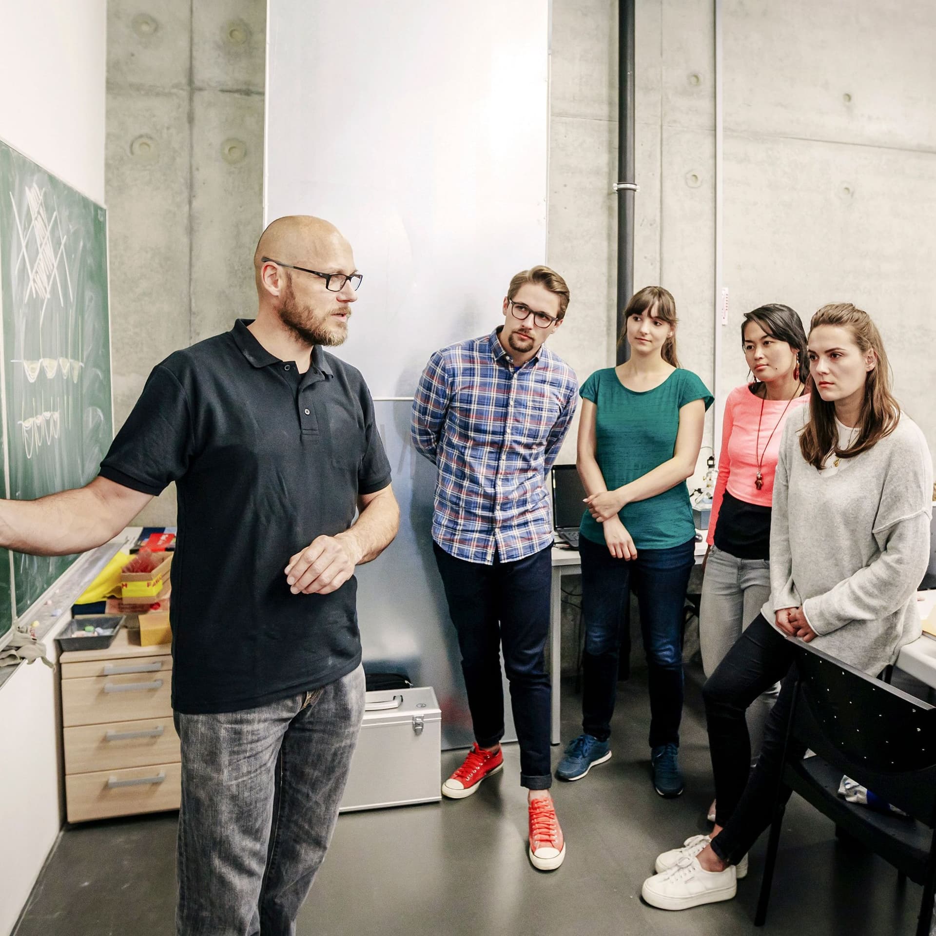 Studenten stehen vor einer Tafel und hören ihrem Lehrenden zu, der eine Kurve an die Tafel gemalt hat.