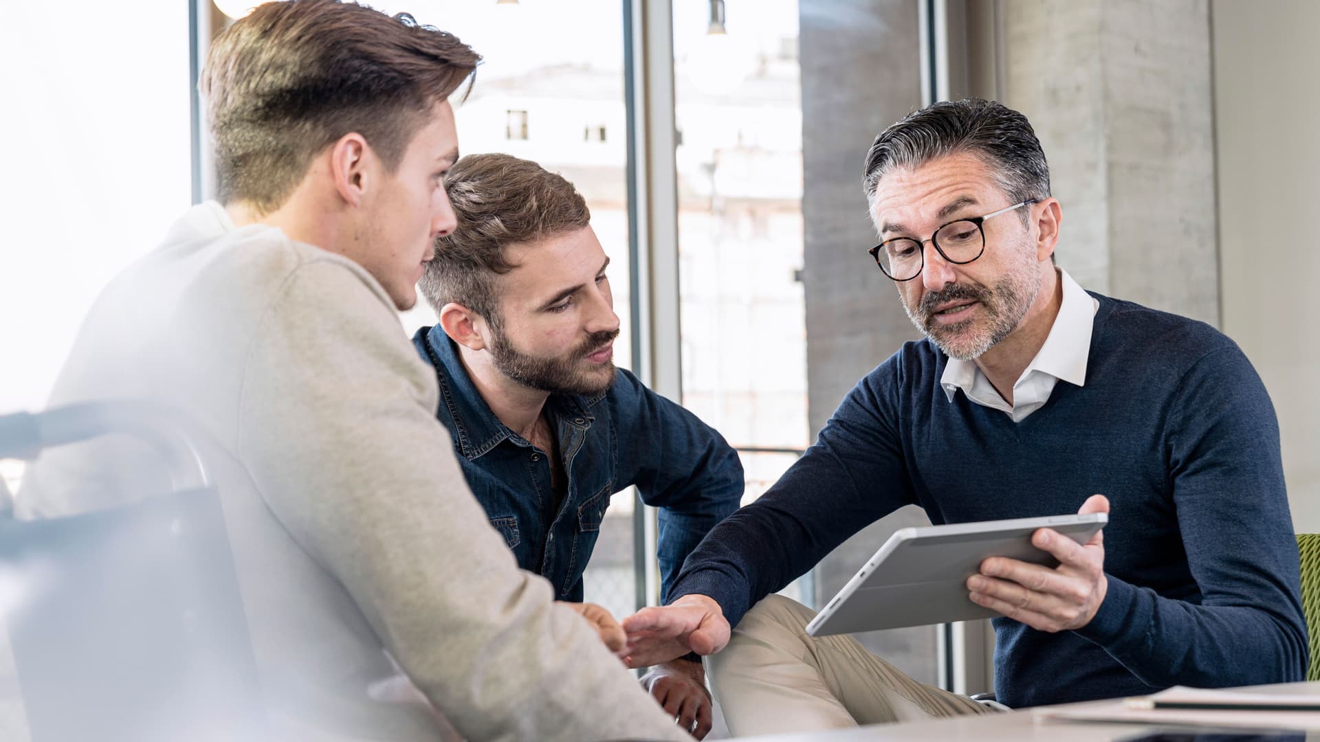 Drei Geschäftsmänner sitzen zusammen in einem Büro und gucken auf ein Tablet.