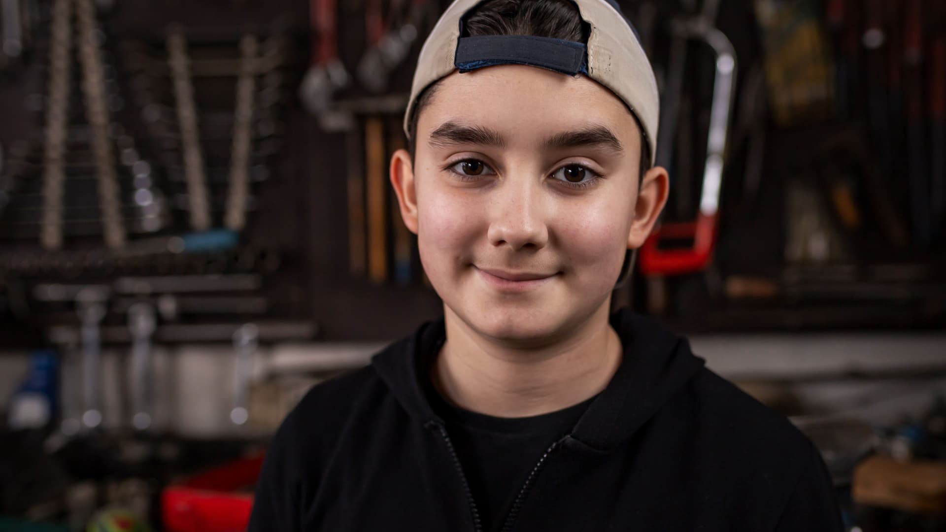 Ein Junge mit weißem Basecap steht in einer Werkstatt. Er lächelt.