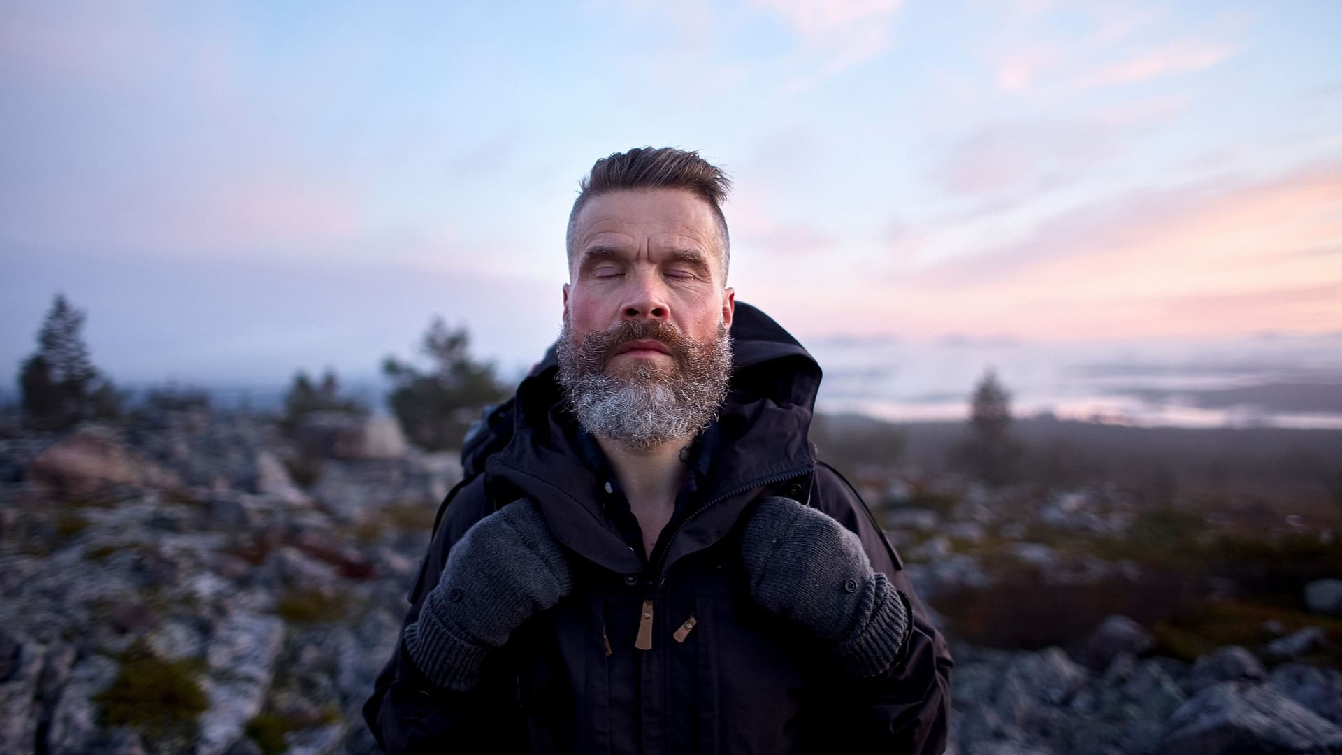 Ein Mann mit grauem Bart steht auf einem Berg mit geschlossenen Augen und genießt den Moment. DIe Sonne ist gerade untergegangen.