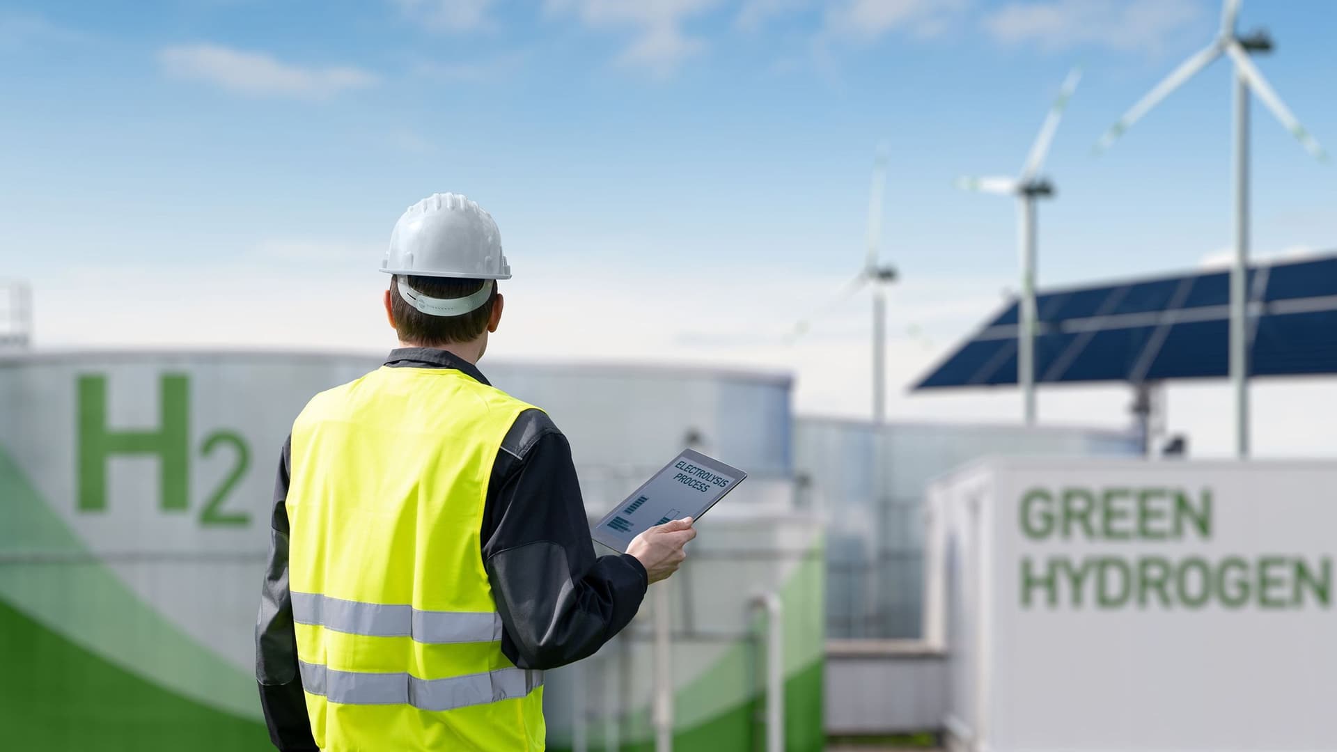 Ein Mann mit Klemmbrett schaut auf einen Wasserstoff-Tank. Im Hintergrund befinden sich Solarpanele und Windräder.