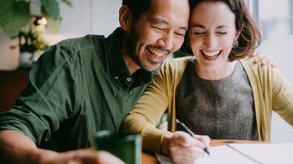 Ein asiatischer Mann und eine Frau sitzen vor Papieren. Sie lachen.