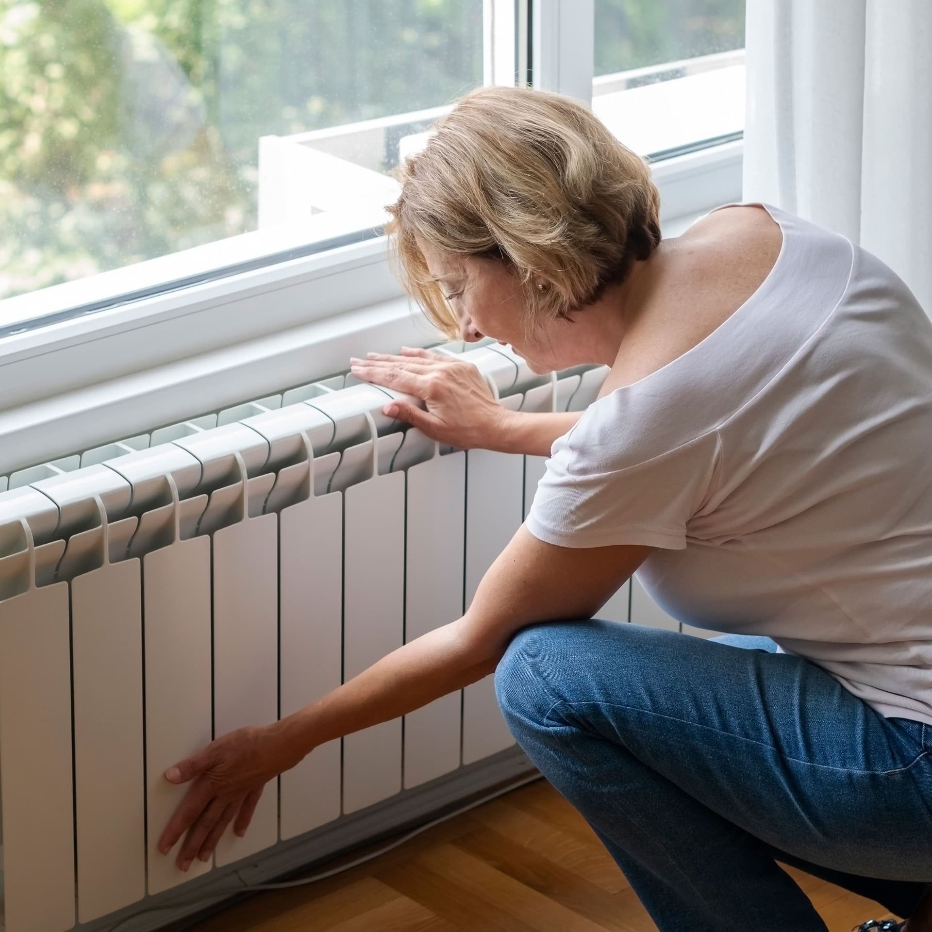 Eine ältere Frau kniet vor ihrer Heizung und fasst sie an.