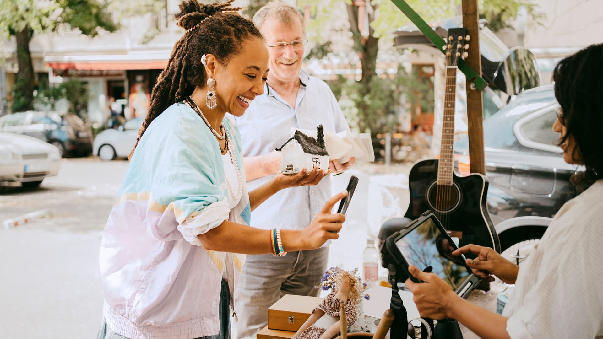 Eine Frau bezahlt auf einem Flohmarkt mit dem Smartphone, während eine Verkäuferin ein Kartenlesegerät hält. Im Hintergrund stehen ein Mann, eine Gitarre und verschiedene Secondhand-Artikel.