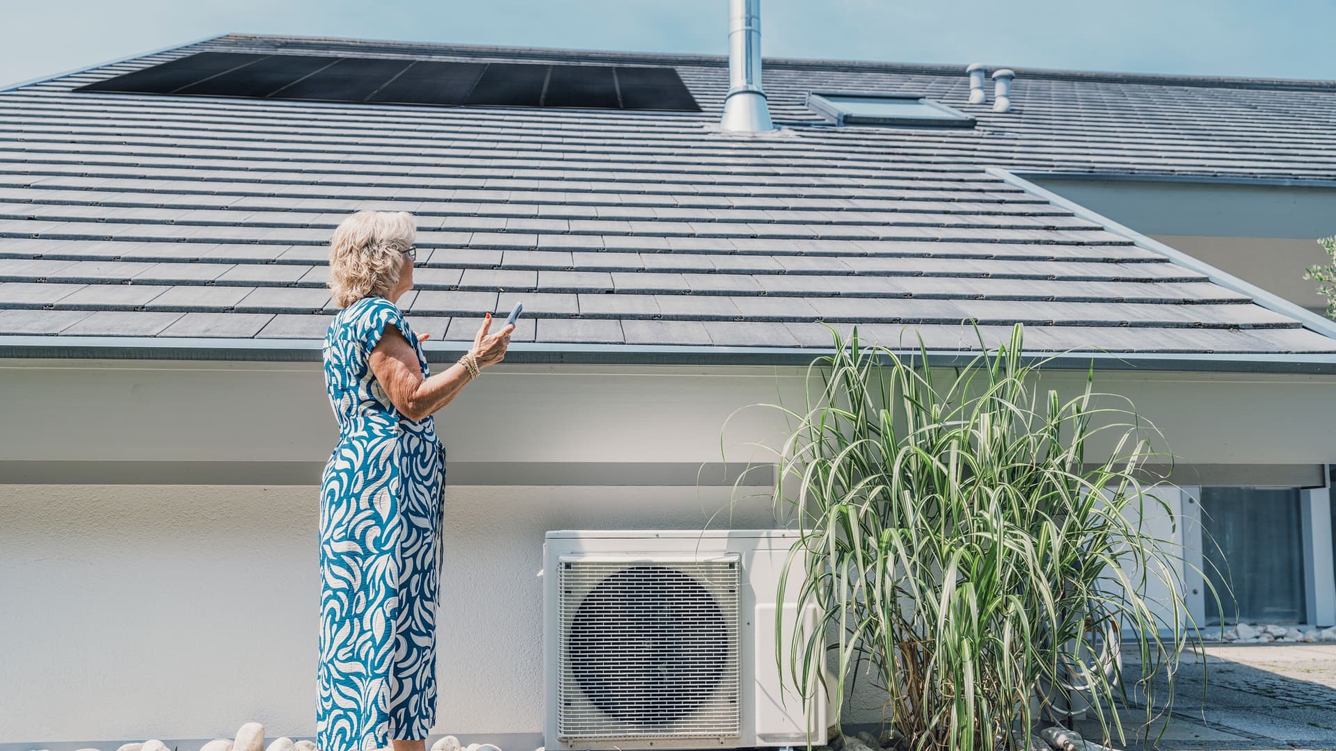 Eine Frau in blau-weißem Kleid steht vor einem Haus mit Solardach, blickt nach oben und hält ein Smartphone in der Hand.
