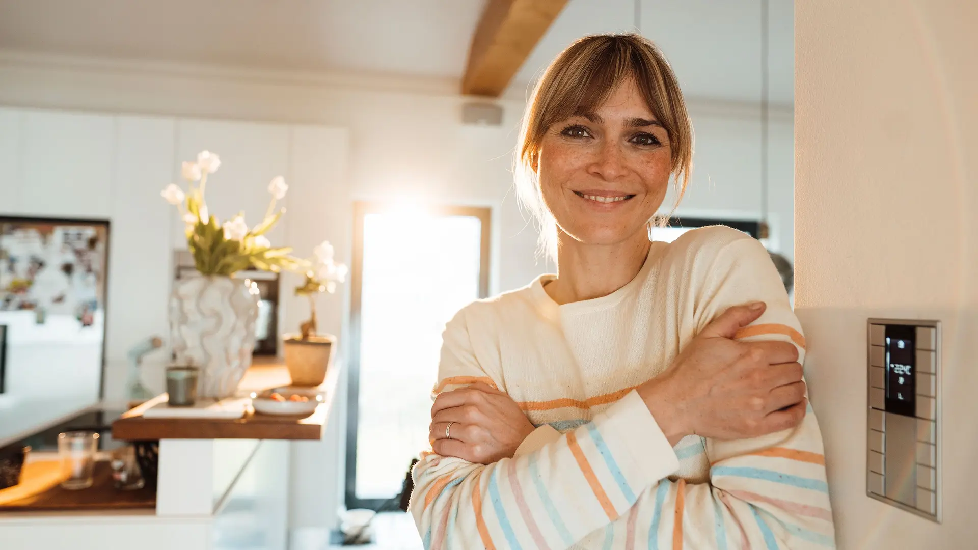Lächelnde Frau steht in einem hellen Wohnraum neben einem Smart-Home-Bedienpanel an der Wand.
