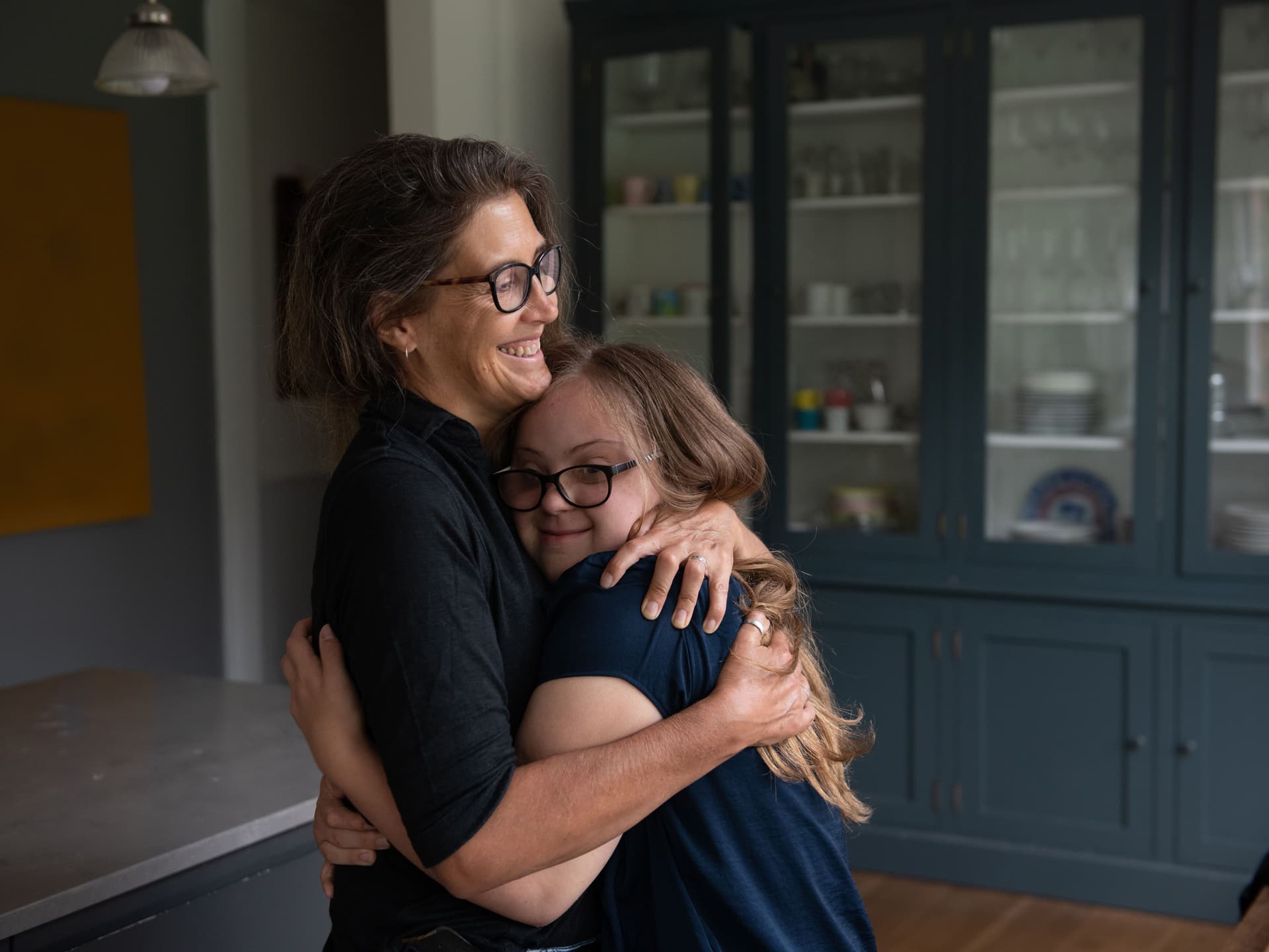 Eine Frau umarmt ihre Tochter mit Downsyndrom. Im Hintergund steht ein dunkelblauer Vitrinenschrank.