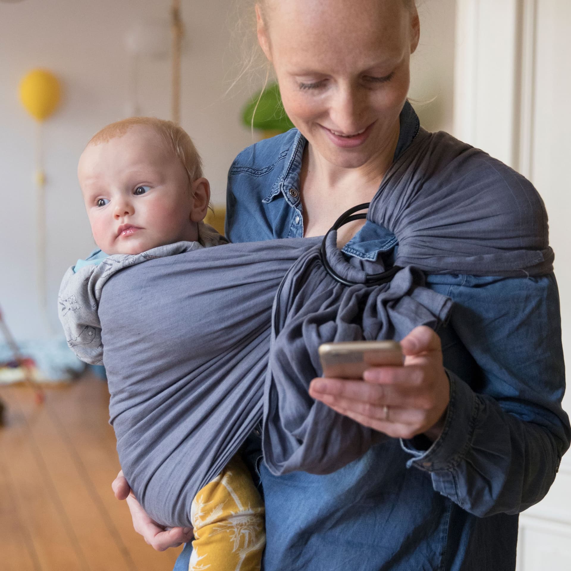 Eine junge Frau mit Kind im Tragetuch schaut auf ihr Handy. Sie lächelt.