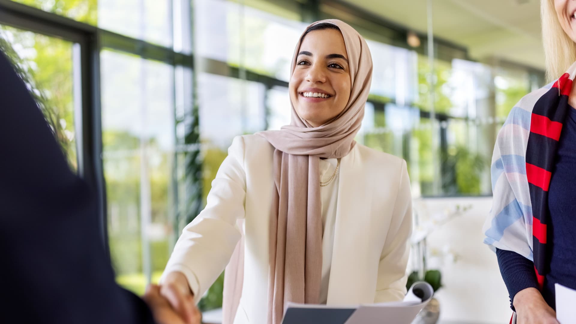Eine junge Frau mit Kopftuch schüttelt einer Person die Hand. Sie hat Unterlagen in der Hand.