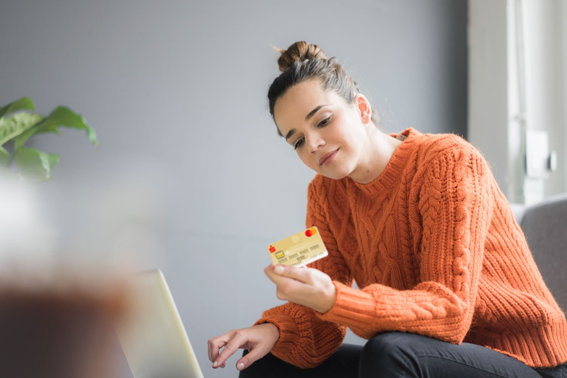Eine junge Frau in orangenen Pulli sitzt vor einem Laptop. In der Hand hat sie eine goldene Kreditkarte. Sie schaut auf die Rückseite der Kreditkarte.