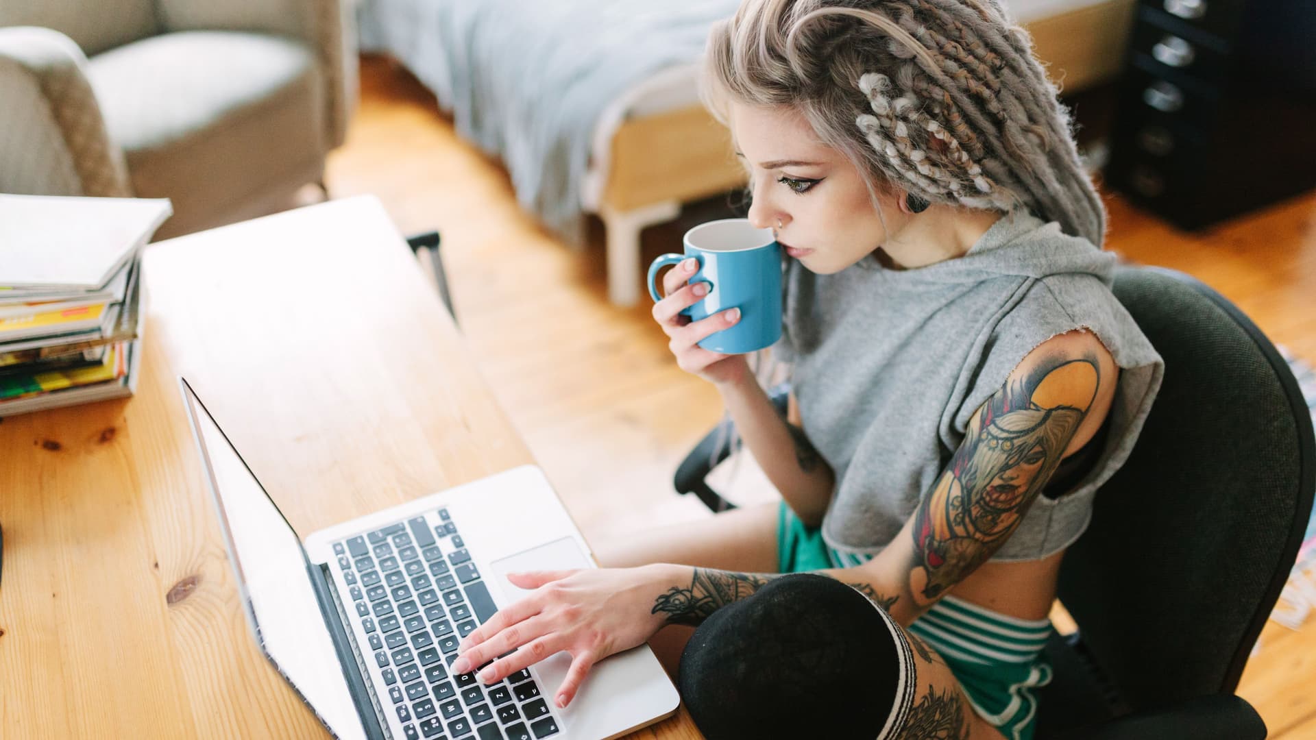 Eine junge tätowierte Frau mit grauen Dreadlocks sitzt konzentriert mit einer Tasse Kaffee vor ihrem Laptop.