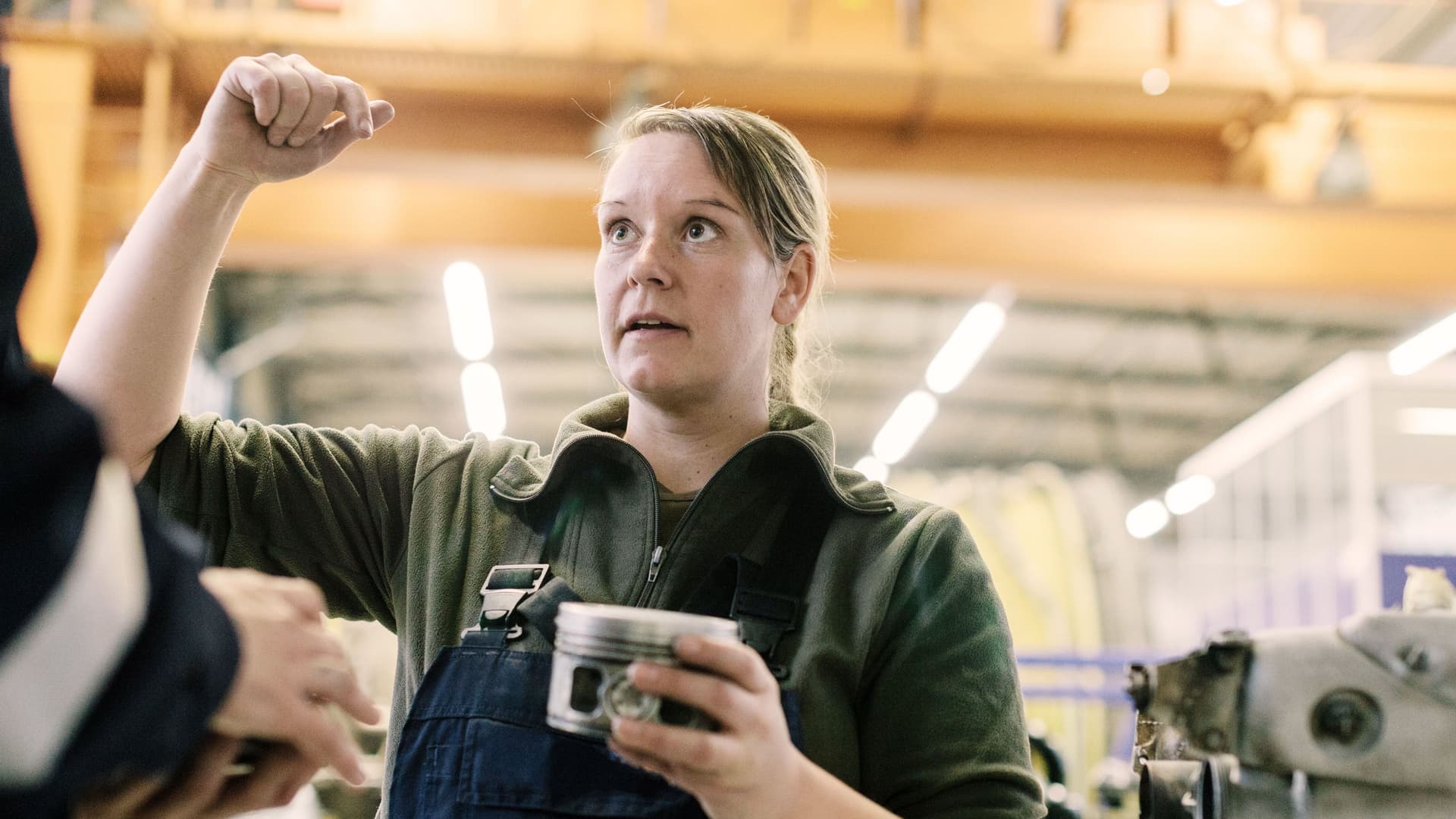 Frau im Blaumann mit Bauteil in der Hand, gestikuliert in einem Gespräch mit einer Person.
