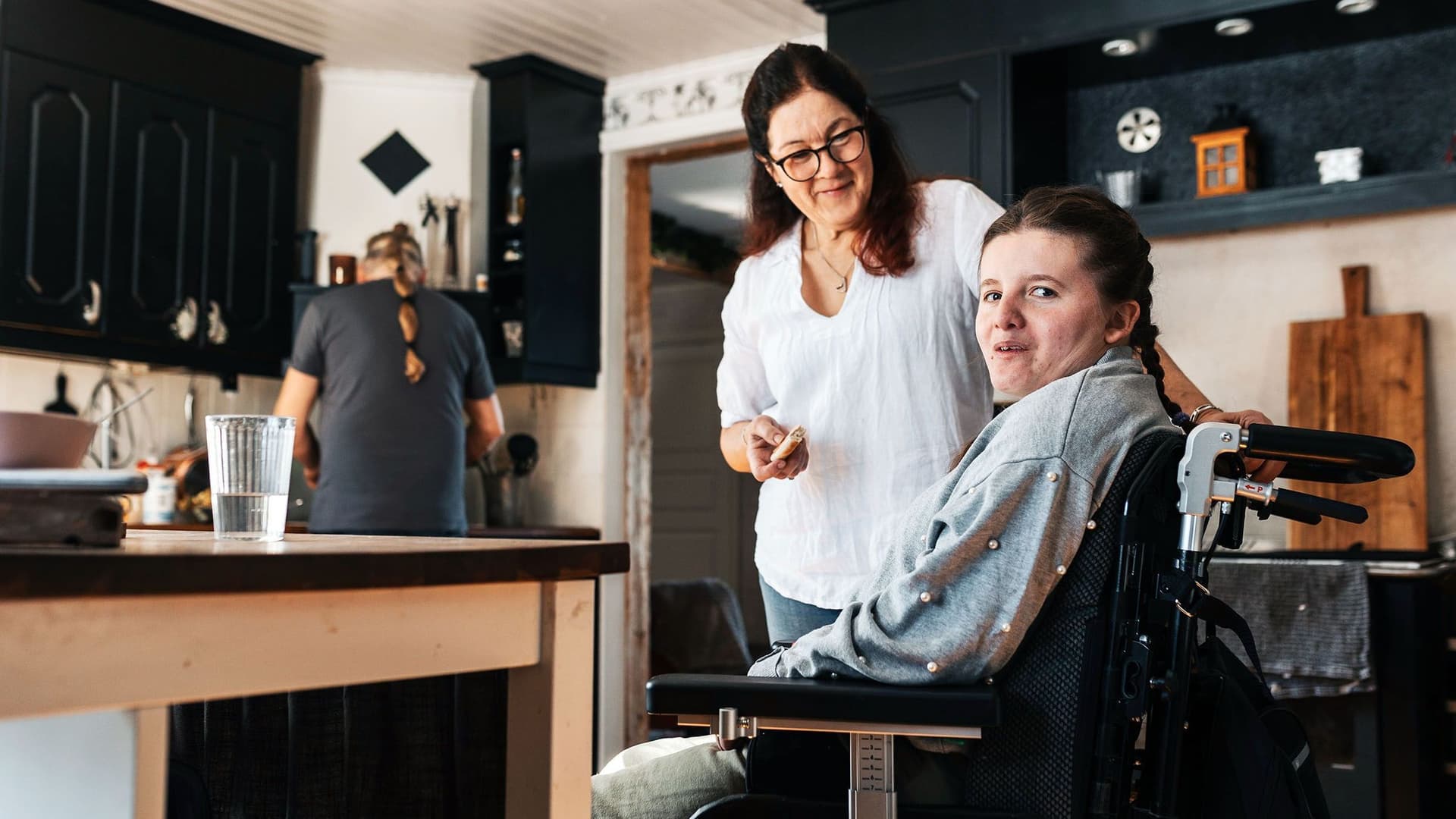 Eine junge Frau im Rollstuhl befindet sich in einer Küche Neben ihr steht eine ältere Frau und lächelt sie an. Im Hintergrund steht eine männliche Person, die in der Küche arbeitet. Die Atmosphäre wirkt familiär und unterstützt