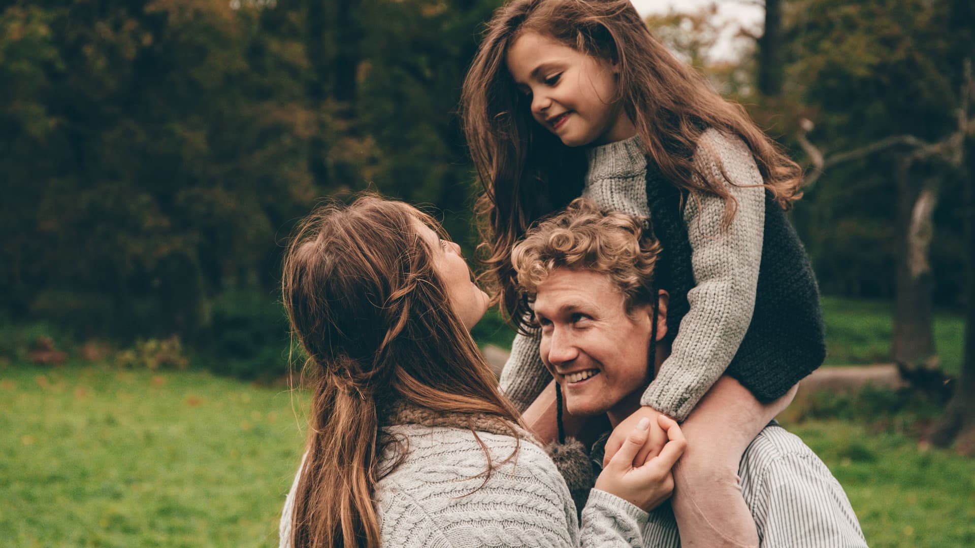 Glückliches Paar mit kleiner Tochter auf den Schultern stehen nah beieinander in der grünen Natur.