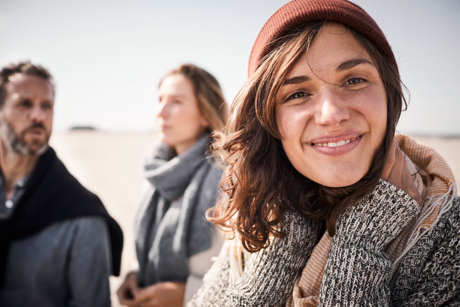 Porträt einer jungen Frau am Strand, die in die Kamera lächelt. Hinter ihr stehen eine Frau und ein Mann.