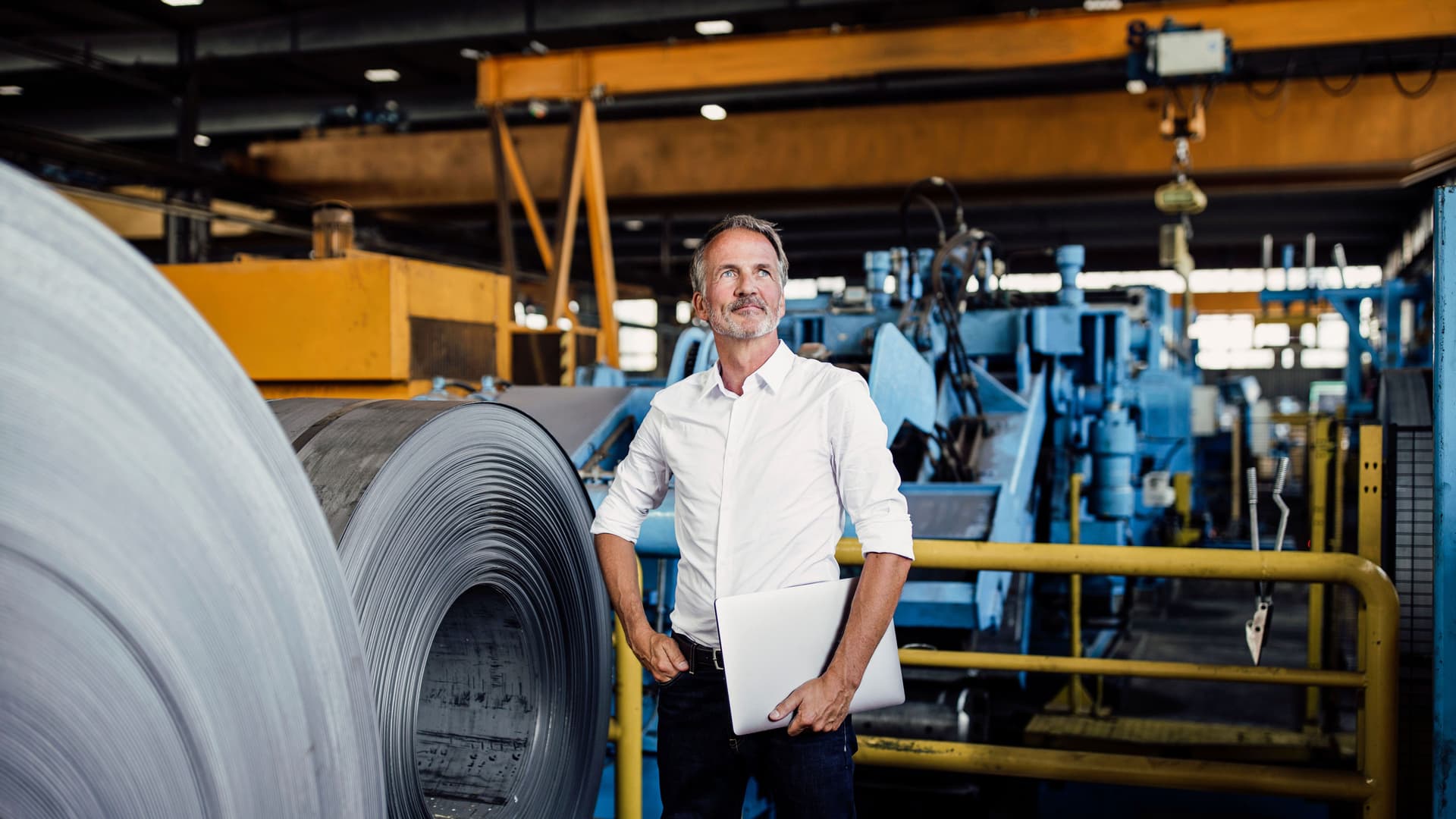 Mann im Hemd steht in einem Lager mit Stahlrollen. Er hat eine Hand in der Hosentasche und hält mit der anderen einen Laptop unterm Arm.