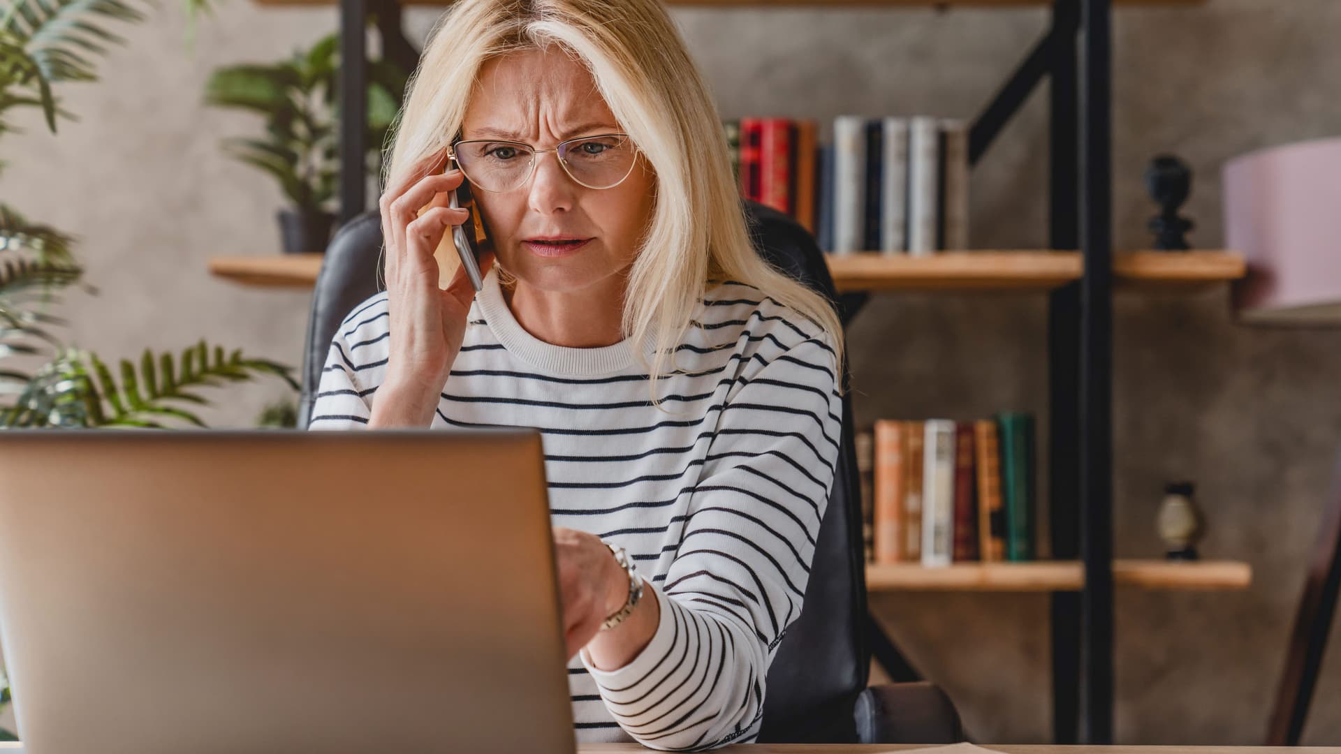Reife Frau mit Streifenpulli sieht gestresst aus, während sie einen Laptop benutzt und mit einem Smartphone telefoniert.