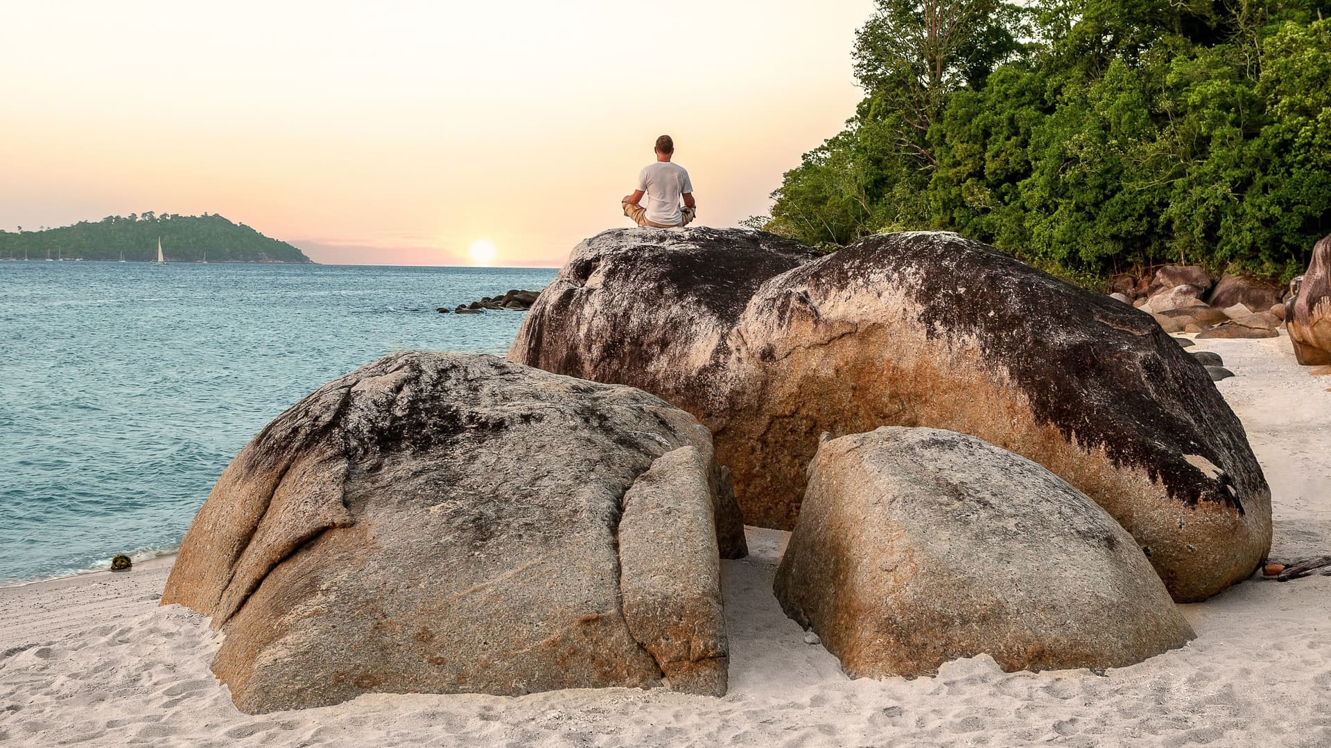 Mann sitzt im Schneidersitz auf Felsen am Strand