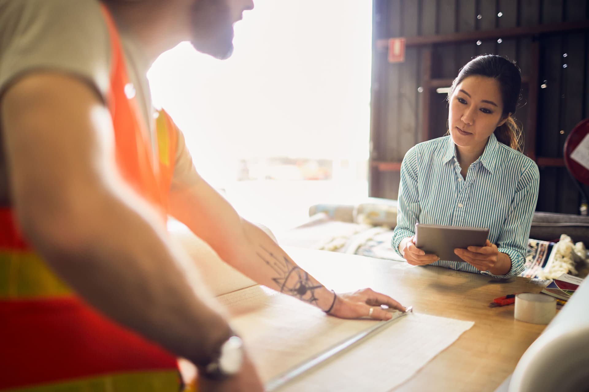 Frau bei einer Konzeptbesprechung in einer Werkstatt. Im Vordergrund sieht man unscharf einen Handwerker, der etwas vermisst.