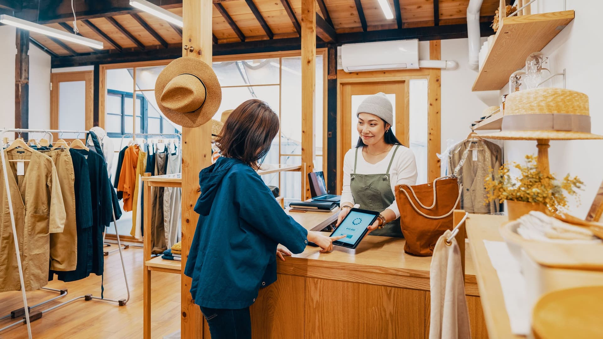 Eine Kundin zahlt digital in einem mit Holz eingerichteten Bekleidungsgeschäft. Die Verkäuferin reicht ihr ein Tablet.