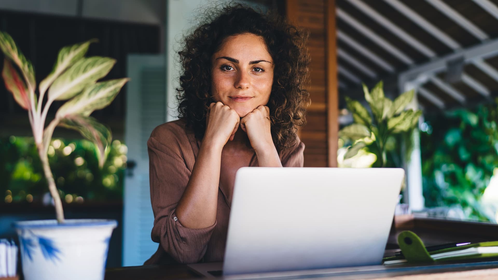 Junge Frau sitzt auf einer Terrasse mit Pflanzen vor einem Laptop. Sie stützt ihren Kopf auf die Hände und lächelt in die Kamera.
