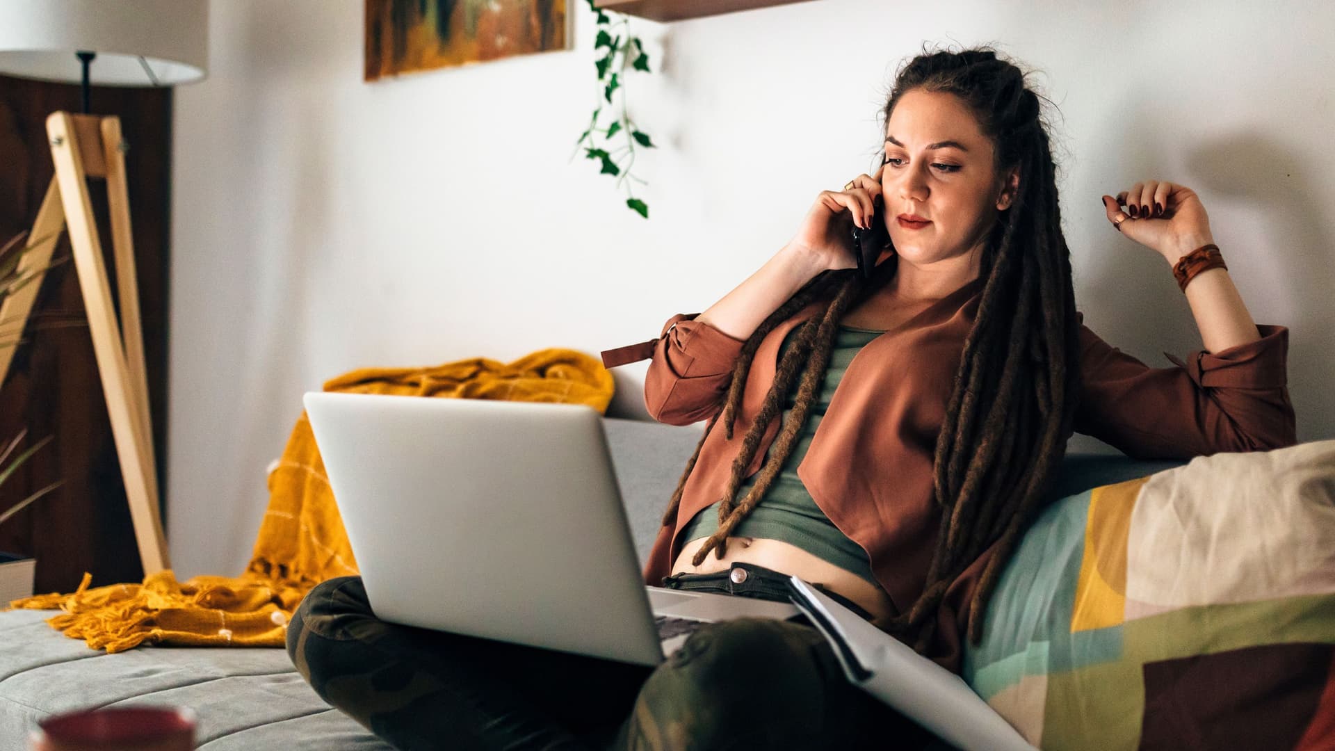 Frau mit langen Dreadlocks arbeitet im Schneidersitz auf dem Sofa. Ihr Laptop steht auf dem Schoß währen sie dabei telefoniert.