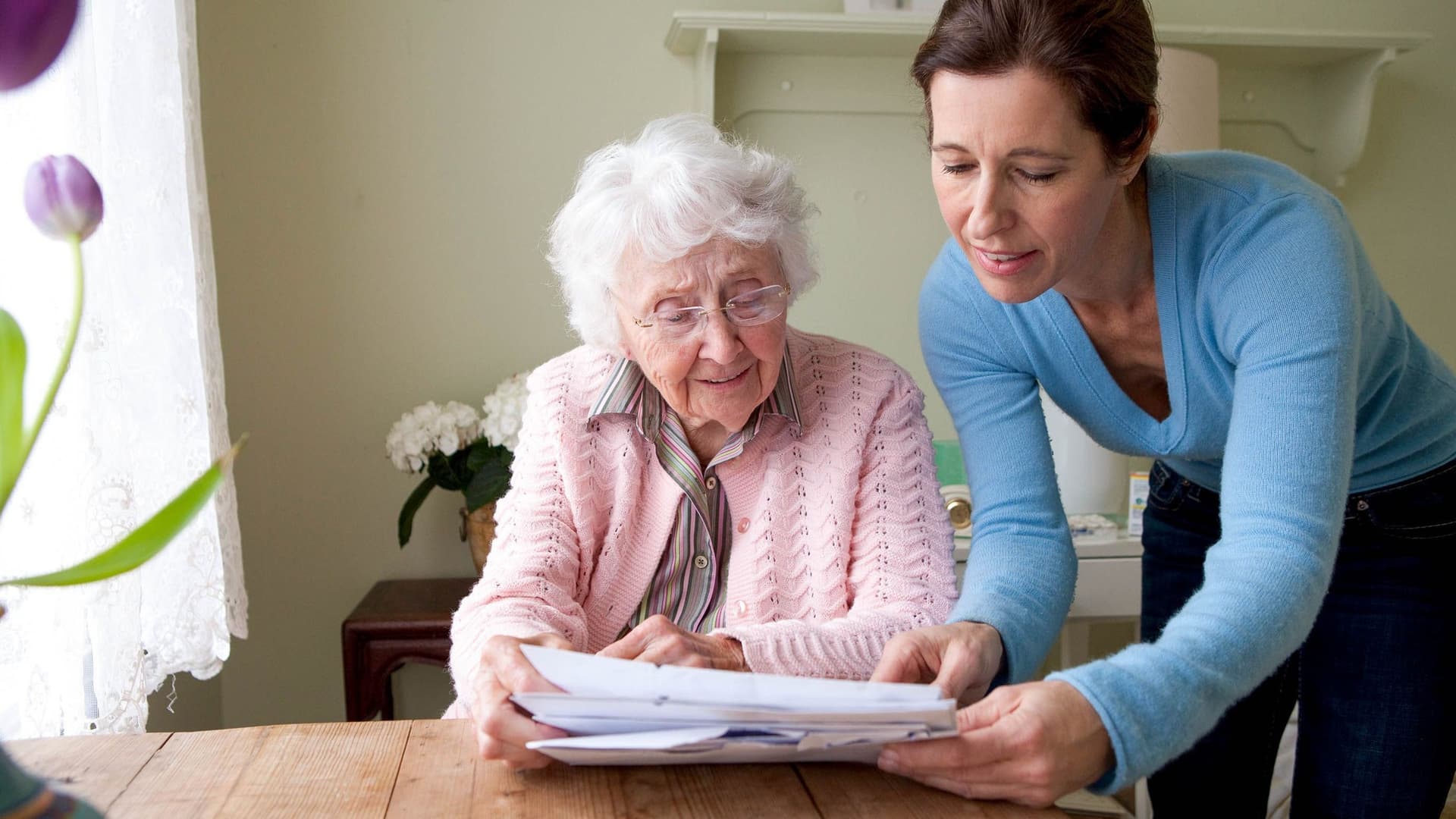 Eine ältere Frau sitzt an einem Tisch und betrachtet zusammen mit einer jüngeren Frau einige Unterlagen. Die ältere Frau trägt eine rosa Strickjacke, die jüngere Frau, die auf die Unterlagen zeigt, einen blauen Pullover.