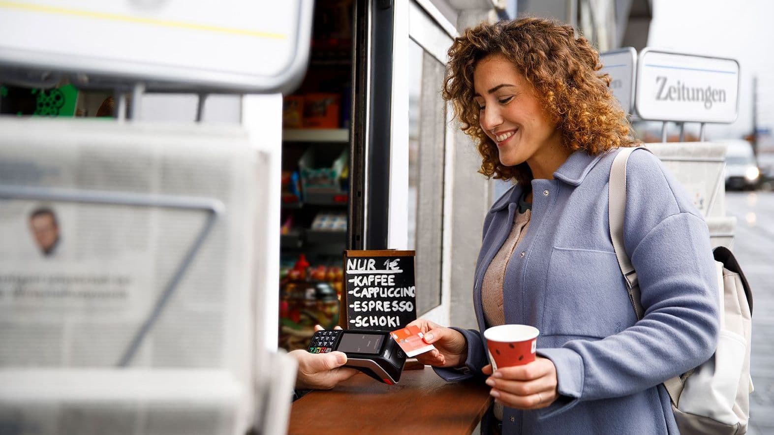 Junge Frau mit dunklen Locken kauft mit der Sparkassenbankkarte ein Heißgetränk an einem Kiosk.